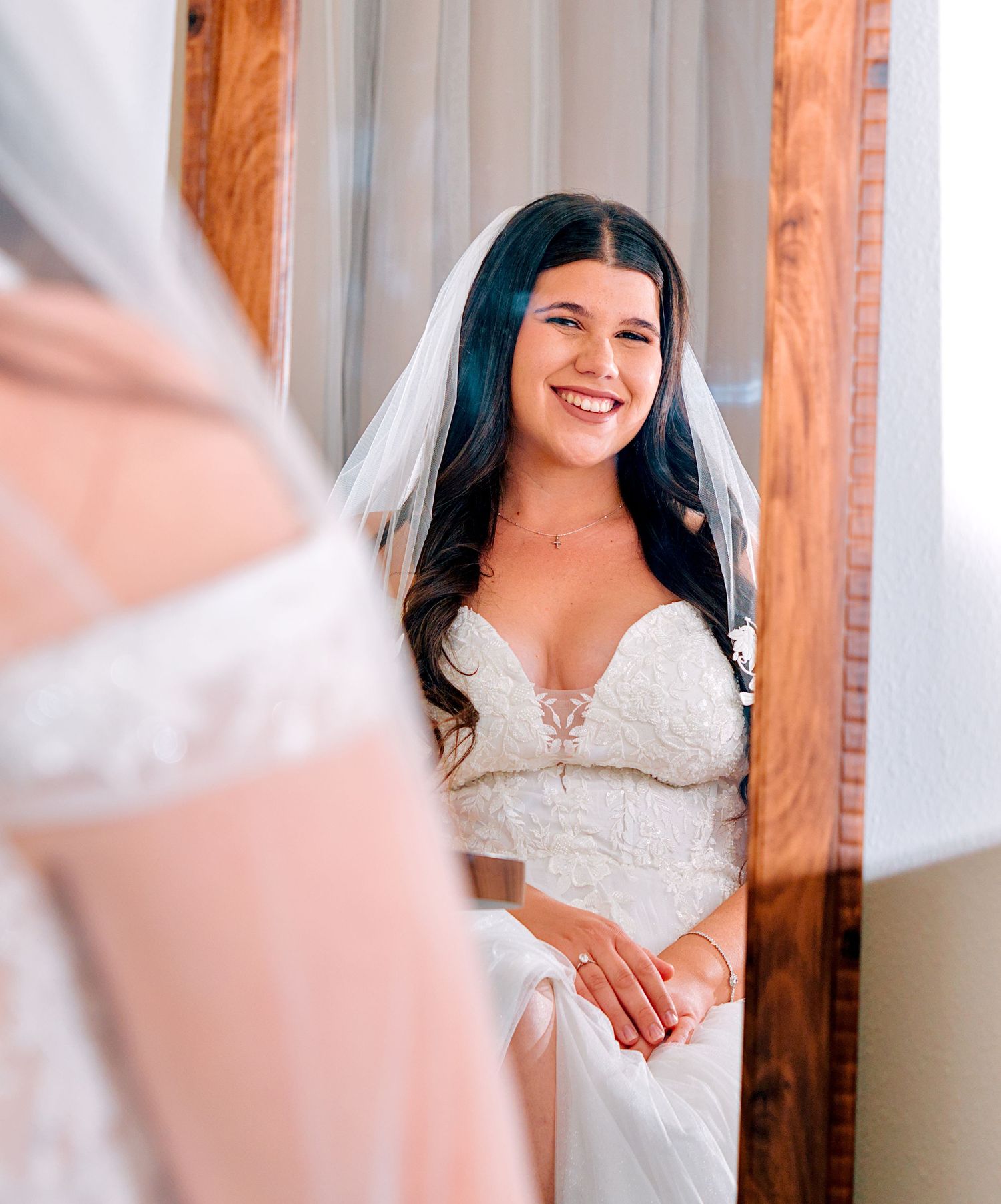 Smiling bride in lace wedding gown and veil sitting before a mirror on her wedding day.