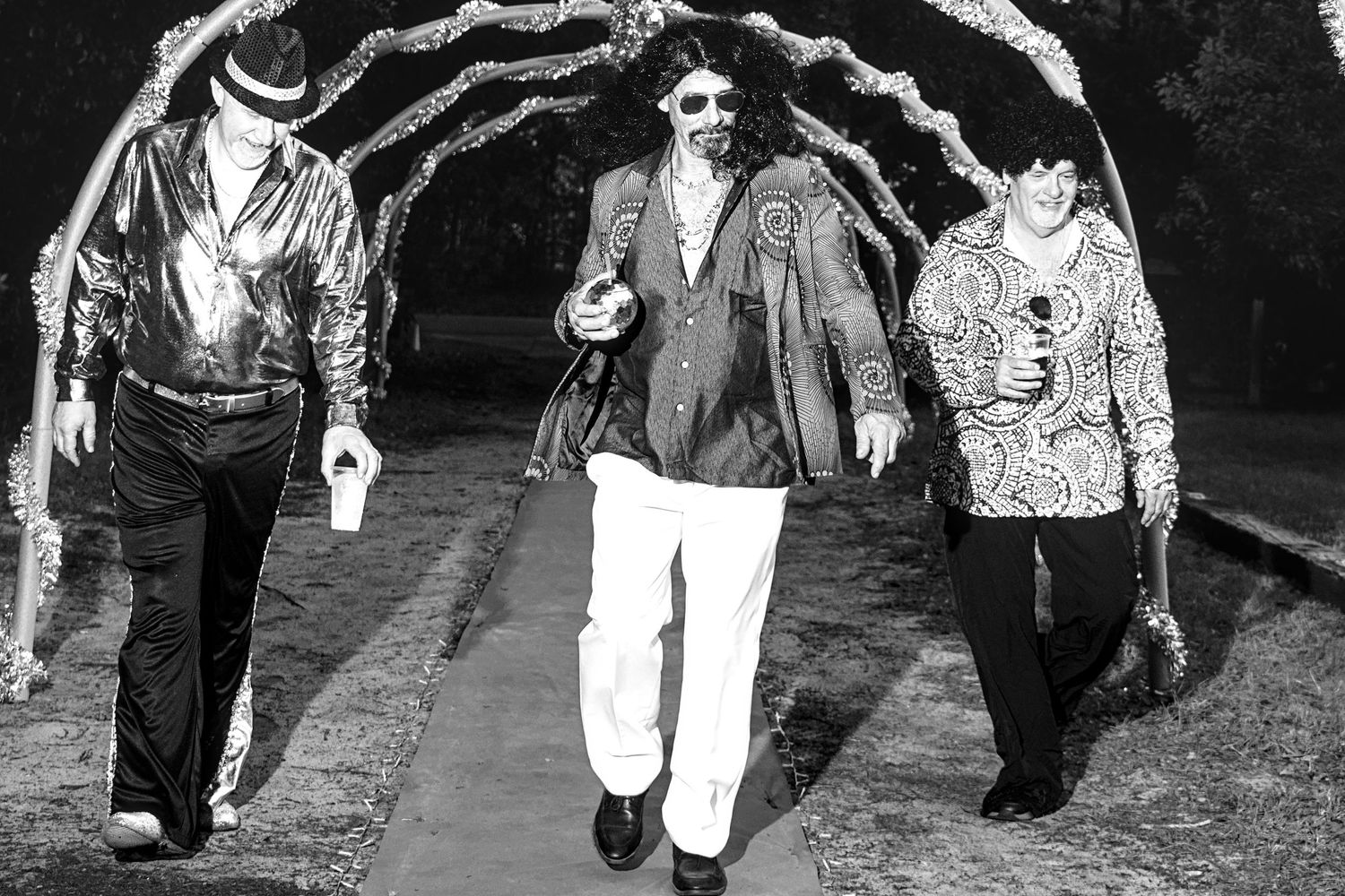 Three people in 1970s disco costumes walk under a decorated arch in a black and white photo.