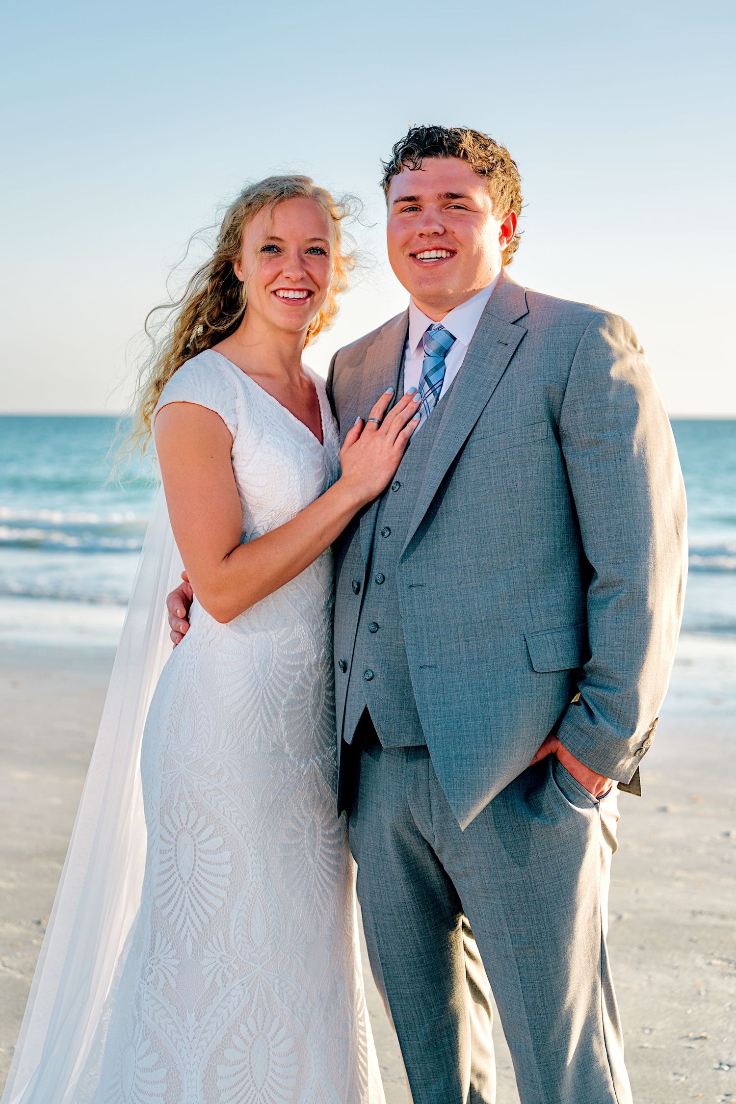 Bride and groom smiling together on a sunny beach, she in a white lace gown, he in a gray suit with blue tie.