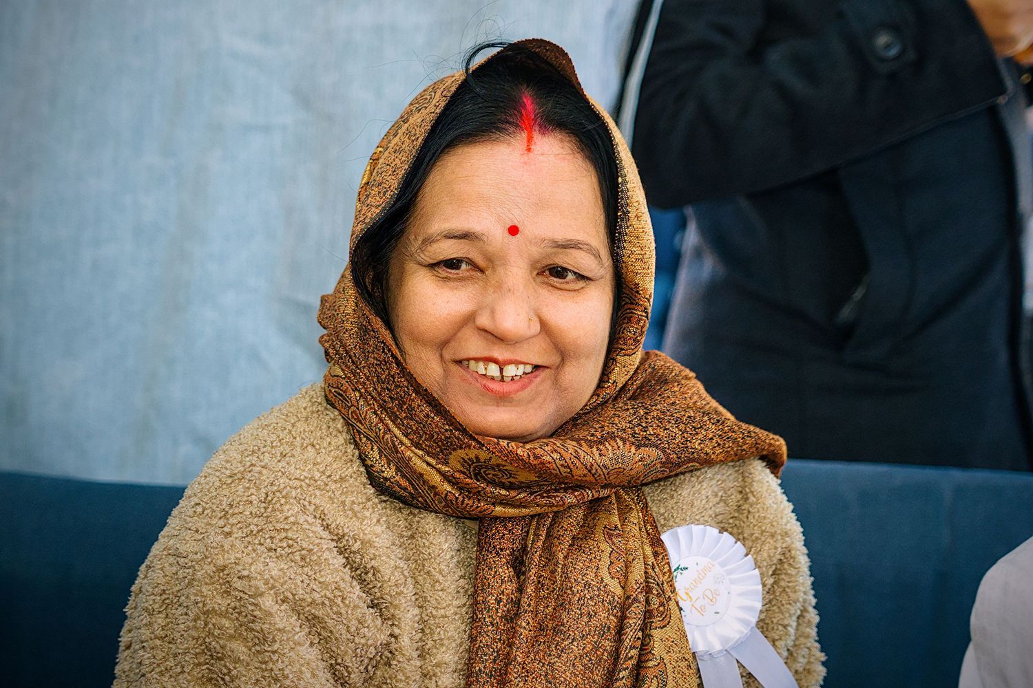A person wearing a brown shawl sits indoors with a warm smile and traditional red bindi marking on their forehead.