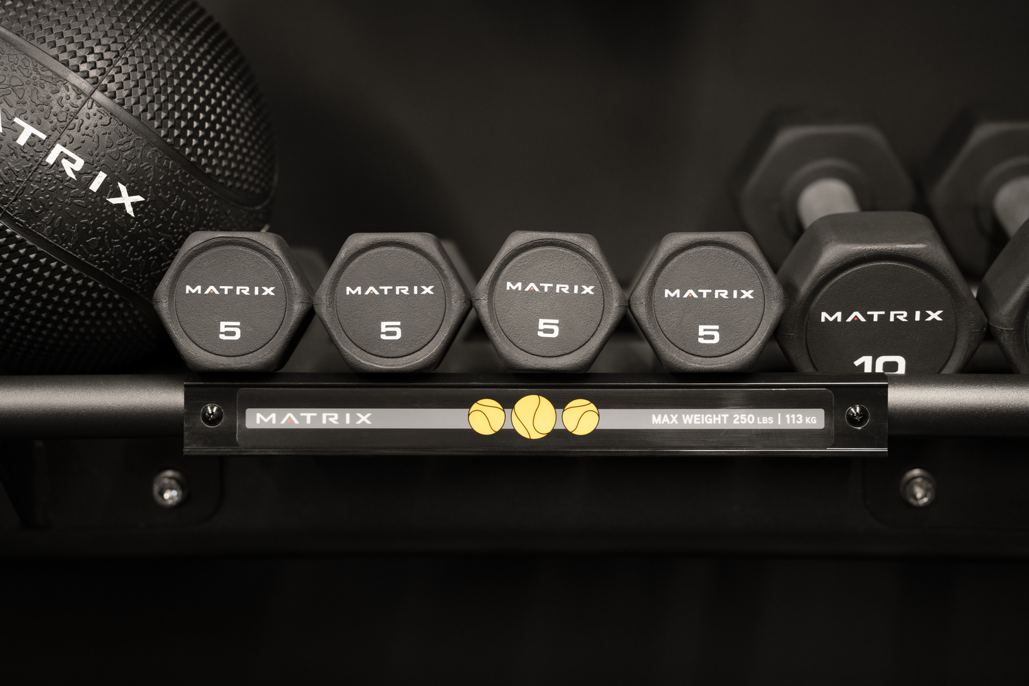 Row of black Matrix 5 lb hexagonal rubber dumbbells on a gym storage rack beside a 10 lb dumbbell and a black medicine ball.
