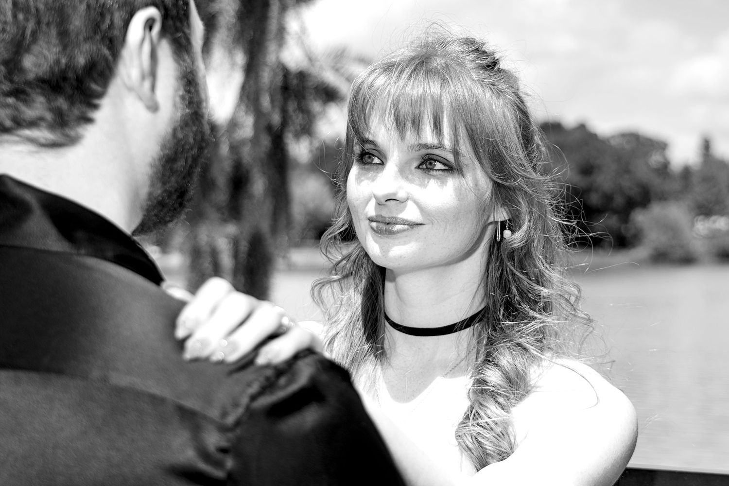 Black and white photo of a young woman with bangs and braided hair smiling at a man outdoors near water.