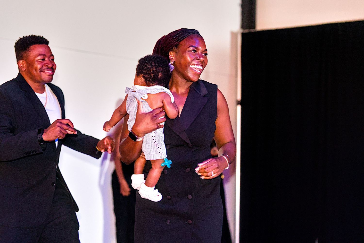 Smiling Black woman in a black dress holds a baby while walking with a man at a celebratory event.