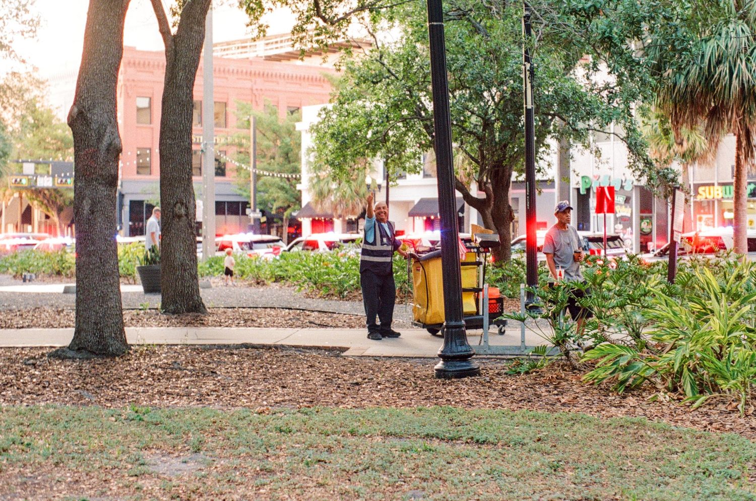 A man stands near a lamppost in a urban park with trees, red flowers, and city buildings in the background.