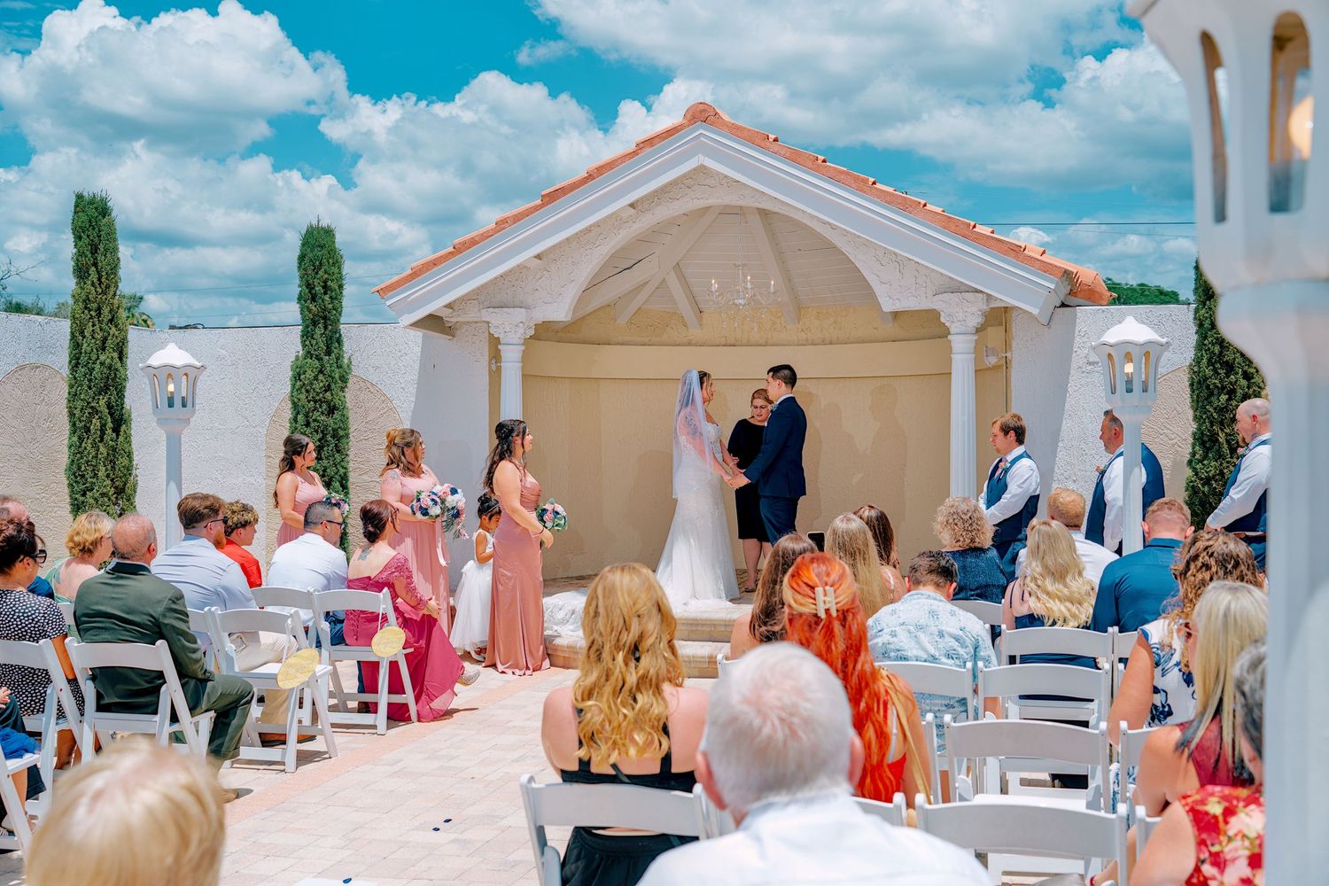 Outdoor wedding ceremony under elegant gazebo with bridal party, guests seated in white chairs, blue sky backdrop.