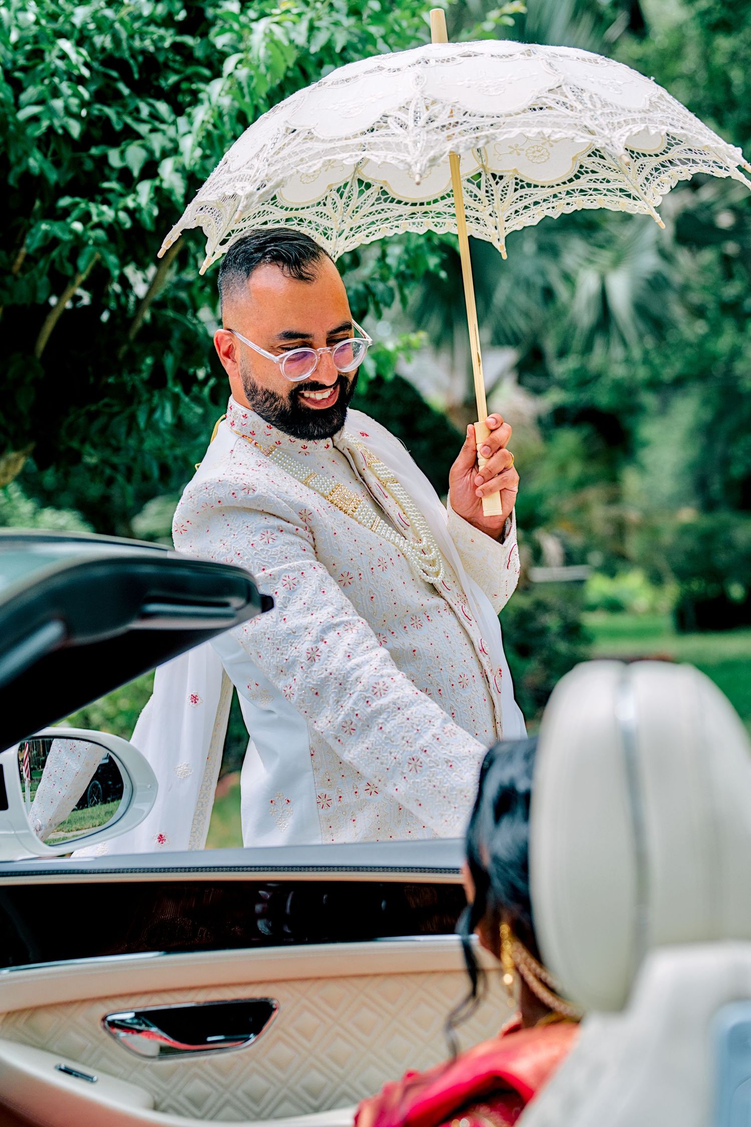 Indian groom in white sherwani holding decorative umbrella beside luxury car.