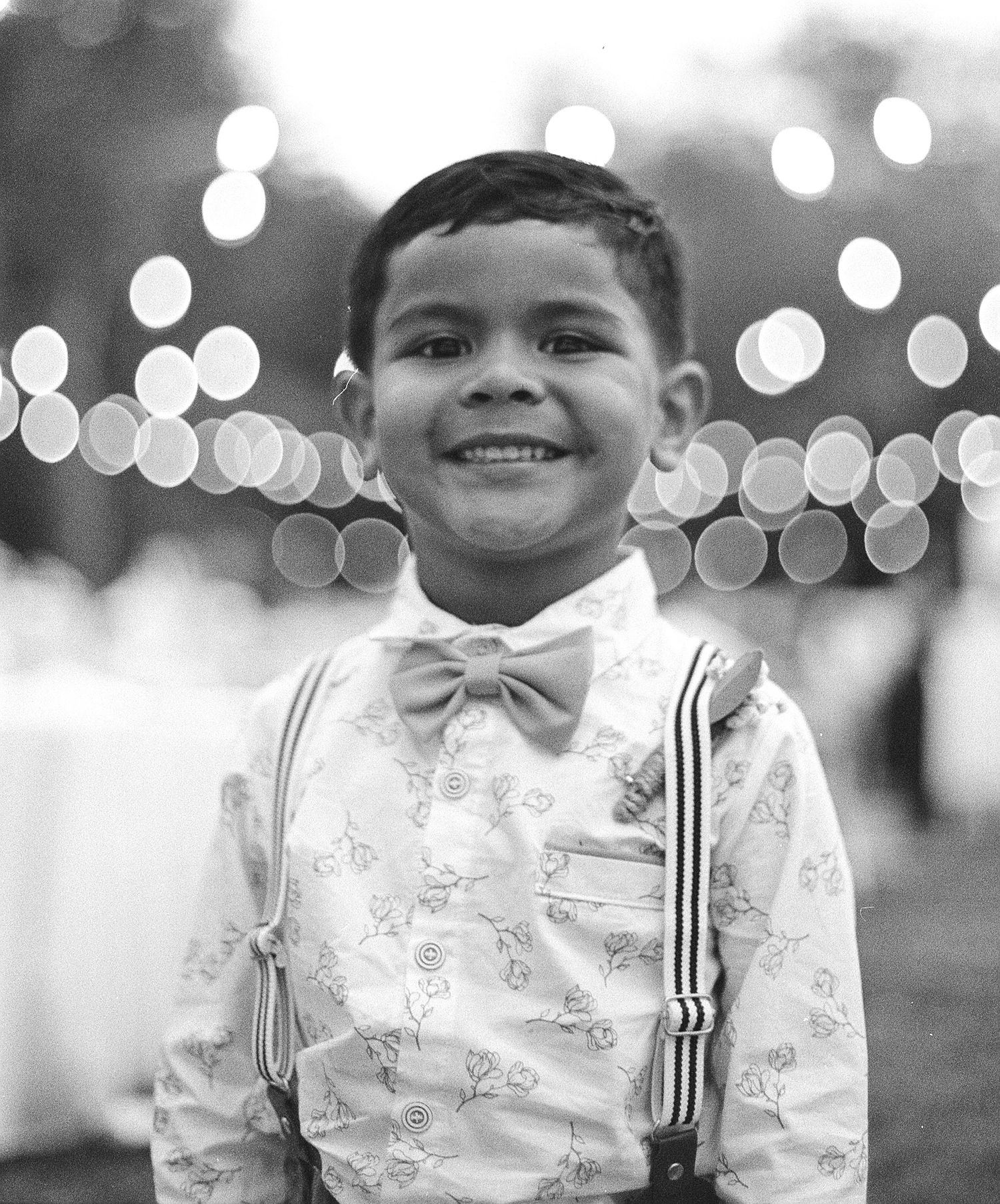 Adorable young boy in bow tie and suspenders smiling at outdoor wedding reception with bokeh lights.