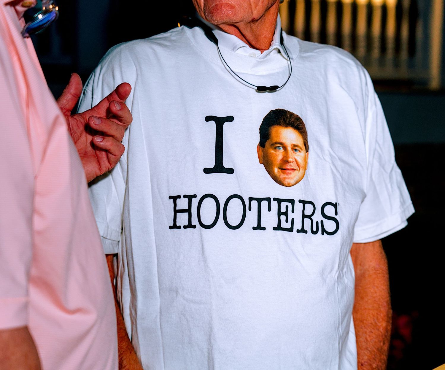 Man wearing a white I Love Hooters parody t-shirt with a face graphic at an indoor event.