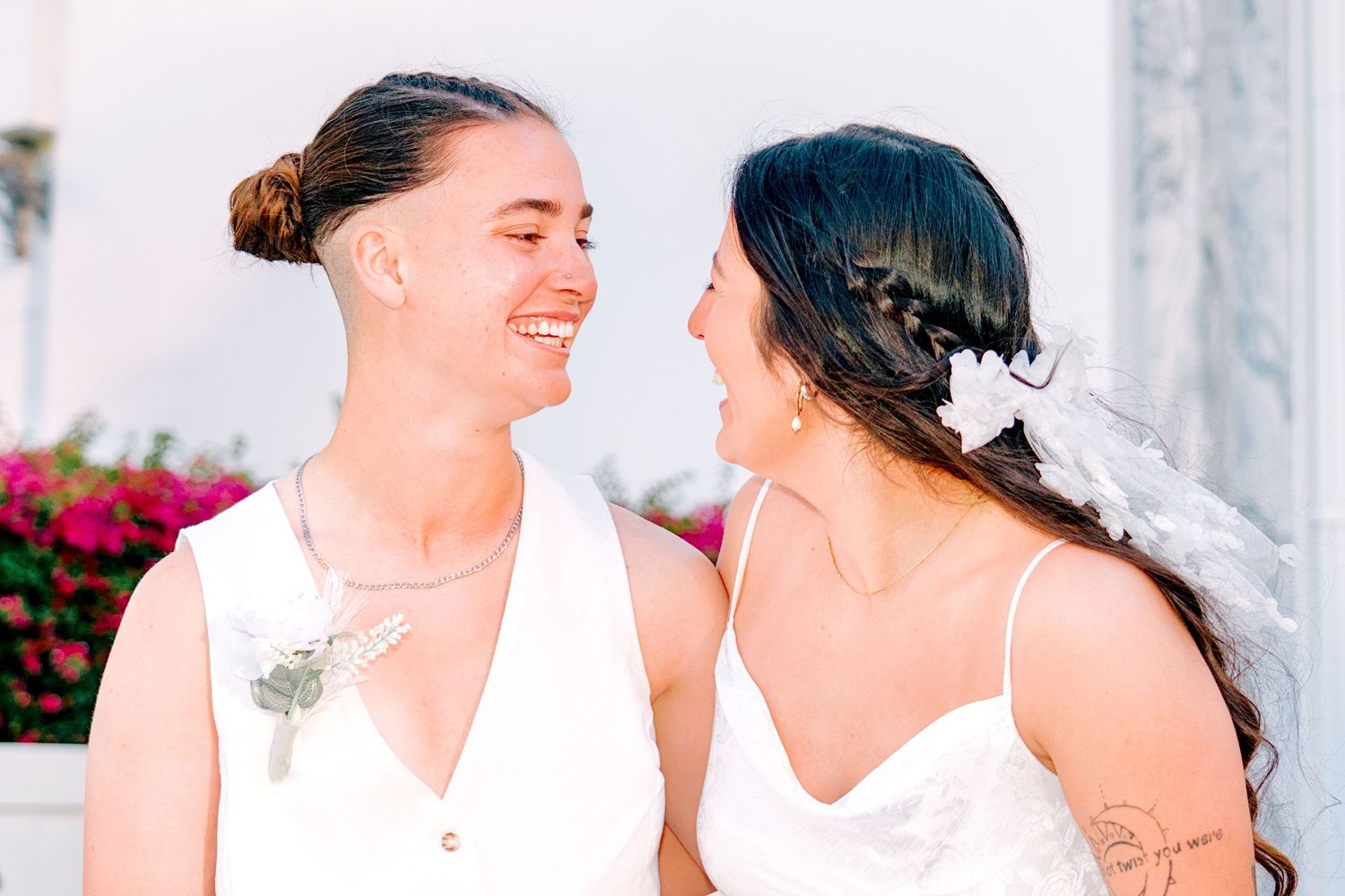 Two brides in white dresses smile at each other during an outdoor wedding ceremony with floral backdrop.