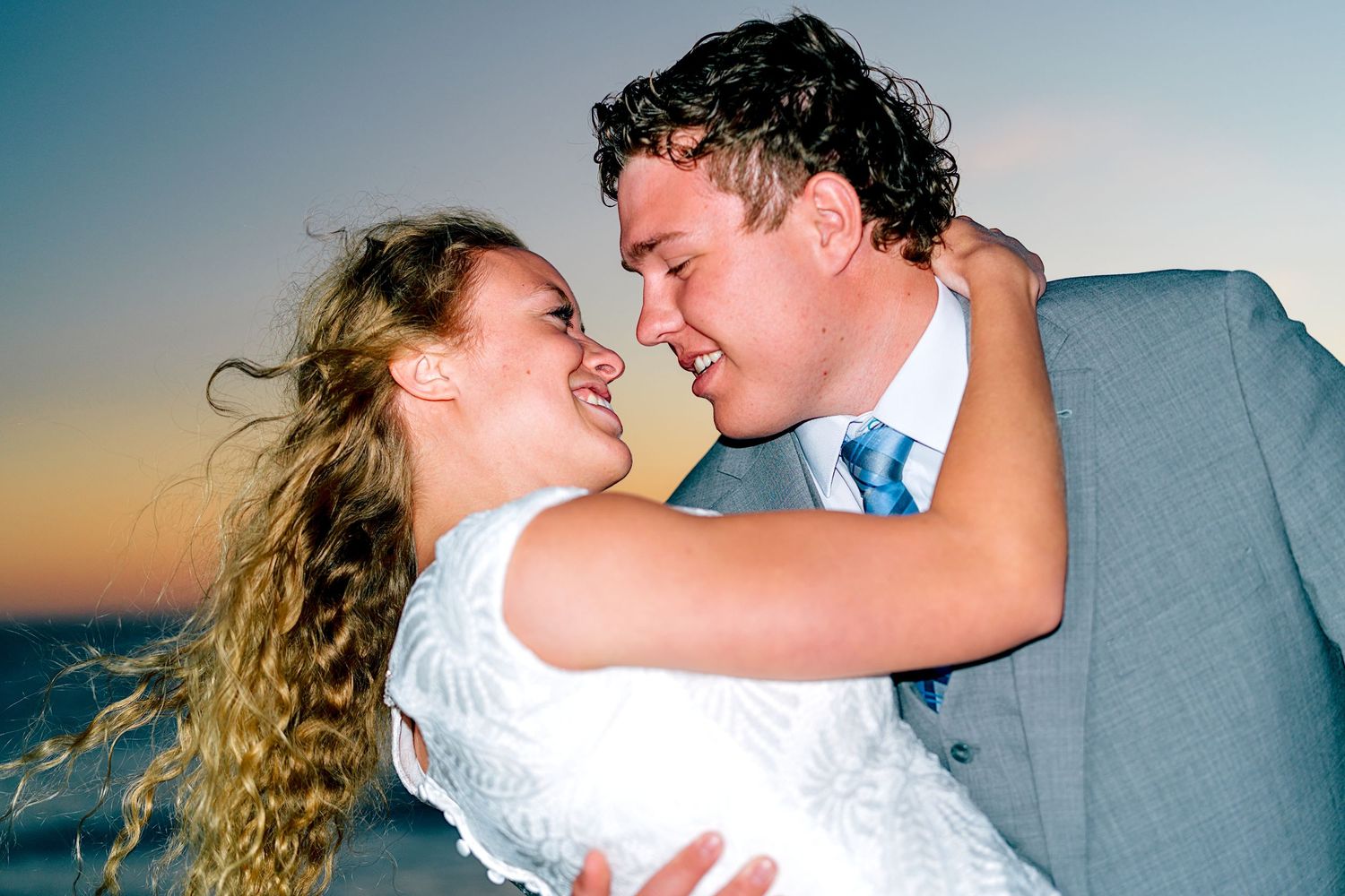 Bride and groom share a romantic moment at beach sunset, smiling and embracing in wedding attire.