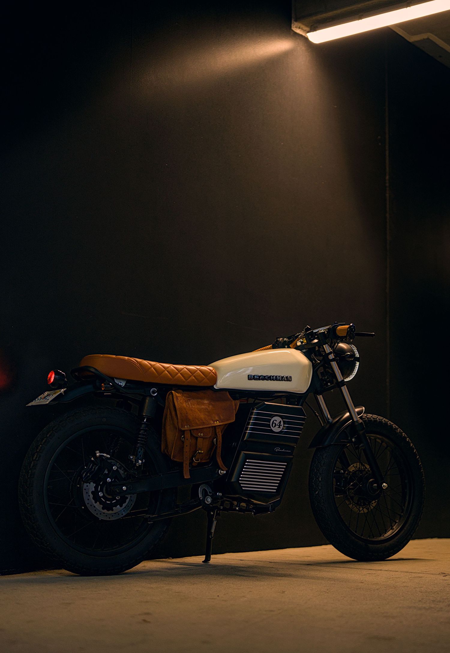 Vintage-style white and tan cafe racer motorcycle parked in a dimly lit garage under a fluorescent light.