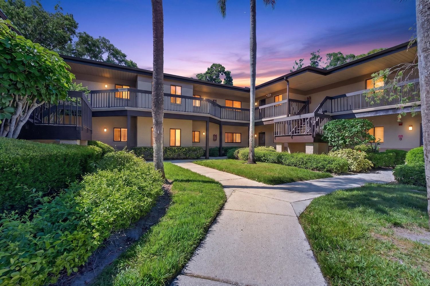 Two-story condo building with lush landscaping, palm trees, and glowing windows at dusk along a winding walkway.