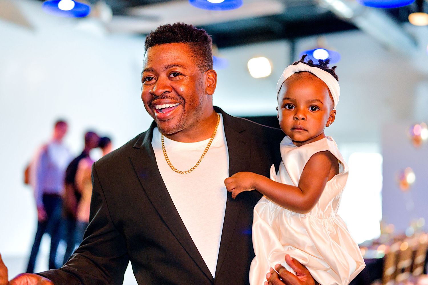 Smiling man in black blazer and gold chain holds a baby girl in white dress at an indoor celebration.