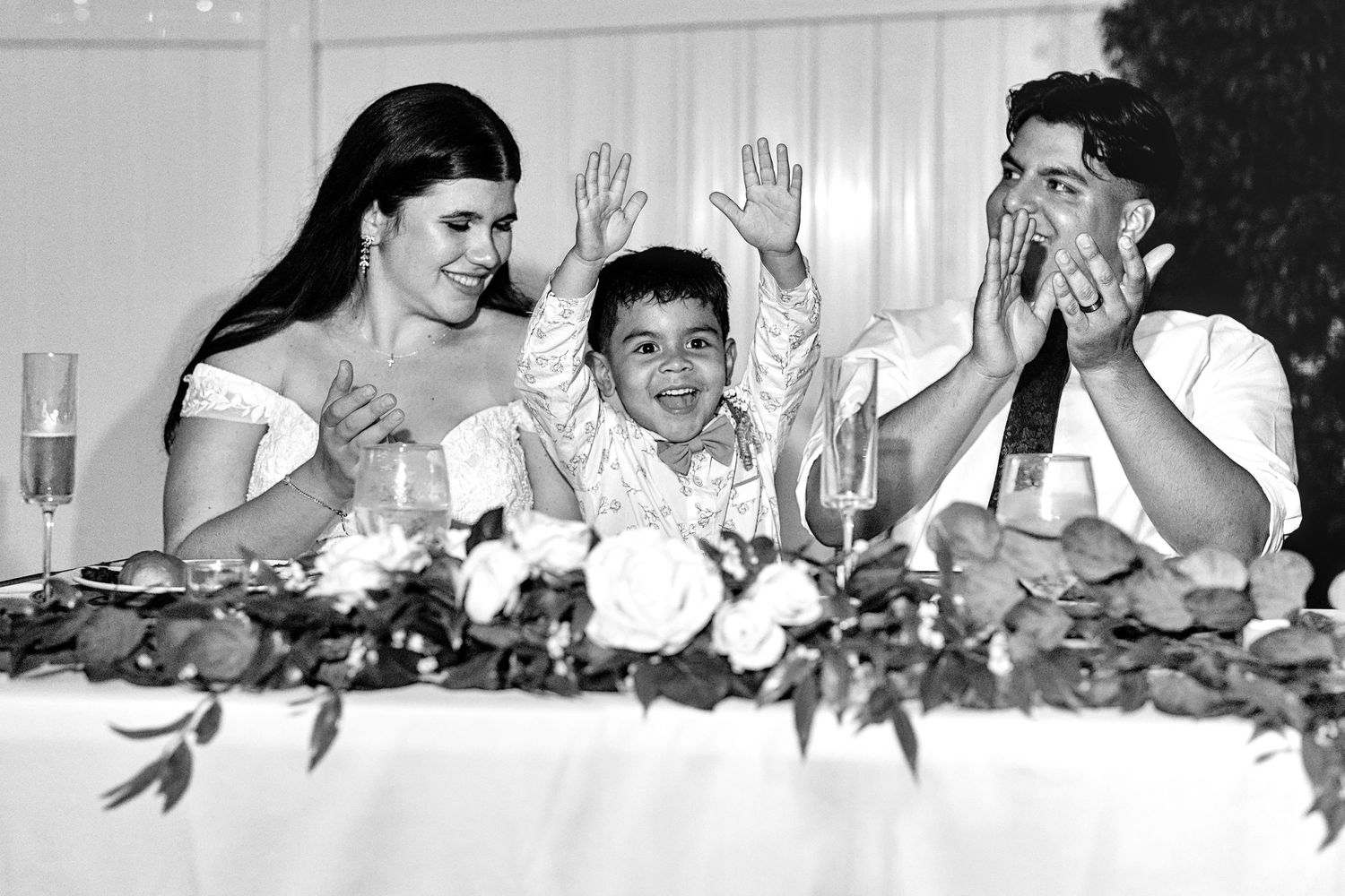 Bride, groom, and child celebrate joyfully at wedding reception table in black and white.