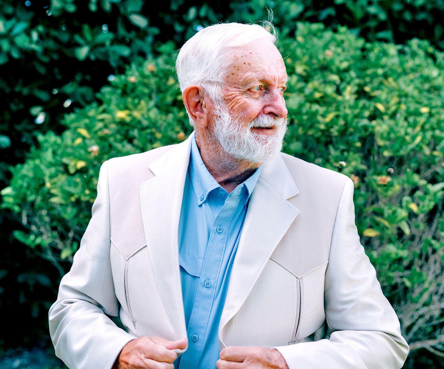 Elderly man with white beard wearing light blazer and blue shirt standing outdoors near green bushes.
