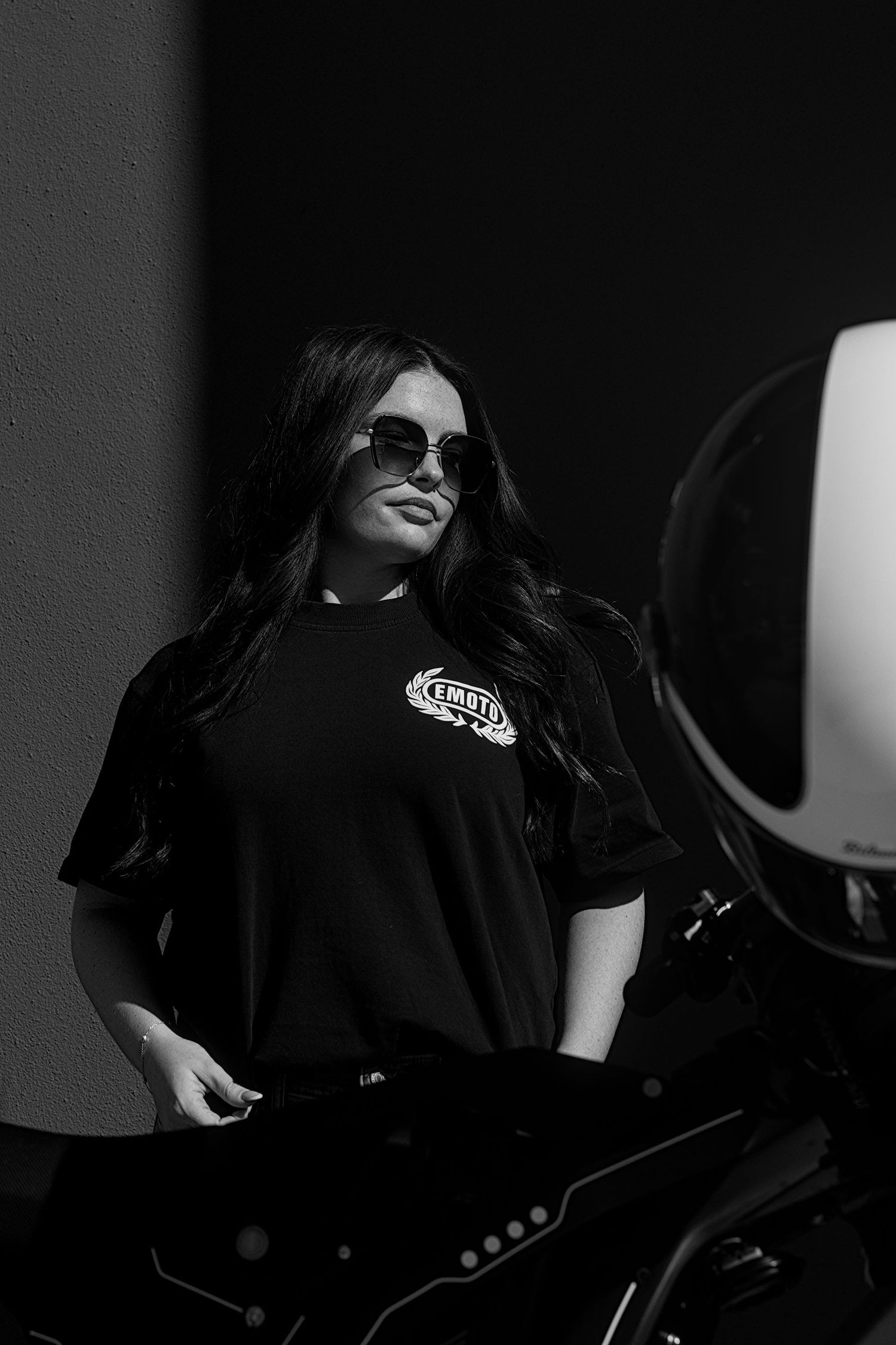 Woman in black graphic tee and sunglasses posing beside a motorcycle in a black and white photo.