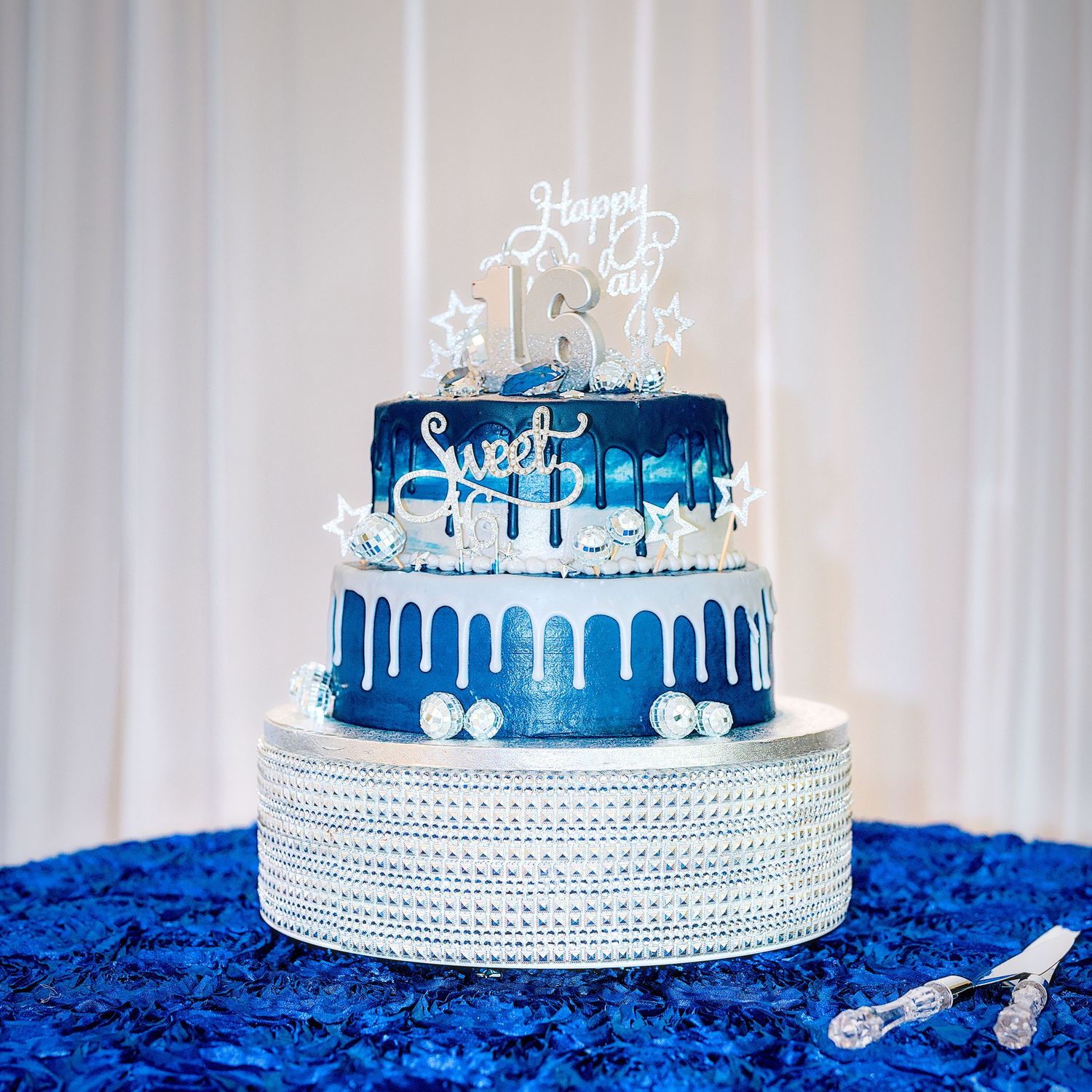 Two-tier blue and silver Sweet 16 birthday cake with drip frosting and Happy 16th topper on rhinestone stand.