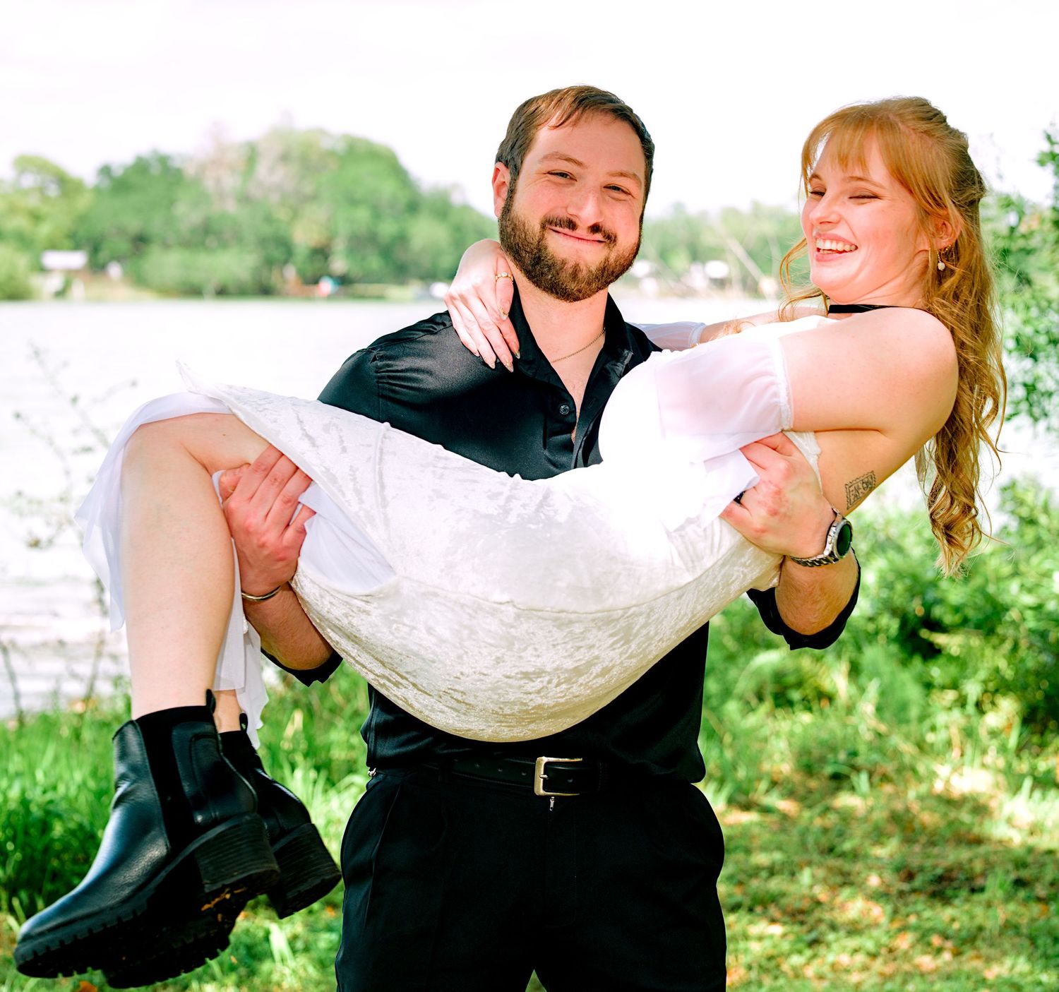 Smiling groom carrying bride in his arms near a scenic lakeside on their wedding day.