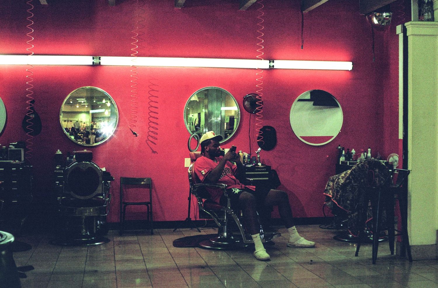Person sitting in a barber chair in a vibrant red barbershop with round mirrors and neon lighting.