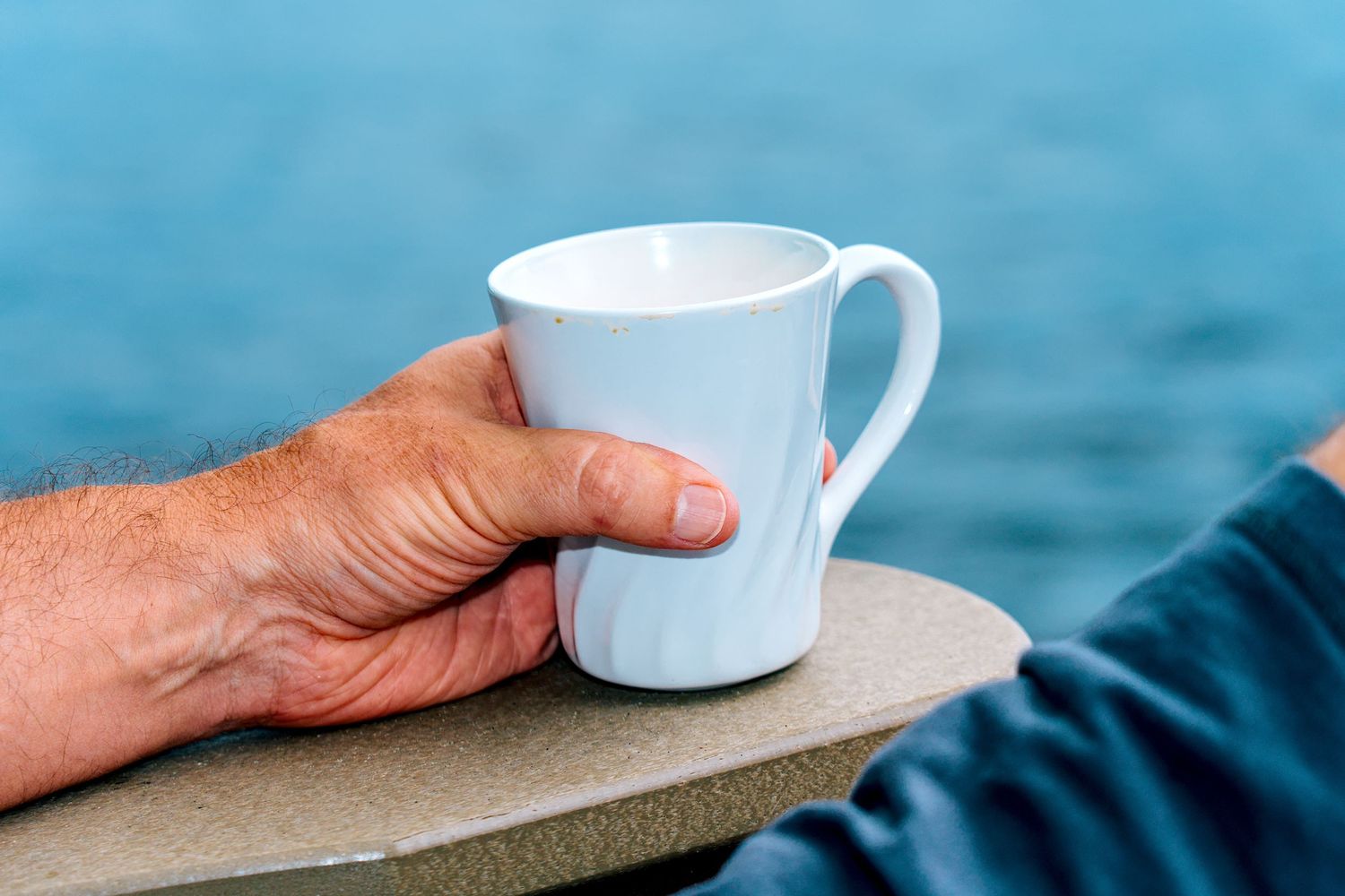 An elderly hand resting on a white ceramic coffee mug on a ledge with a blue ocean background.