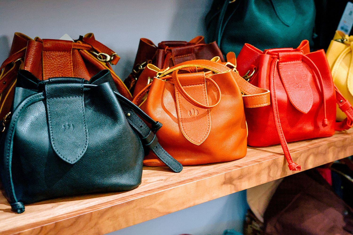 Colorful leather handbags in teal, tan, red, and brown displayed on a wooden shelf in a boutique store.