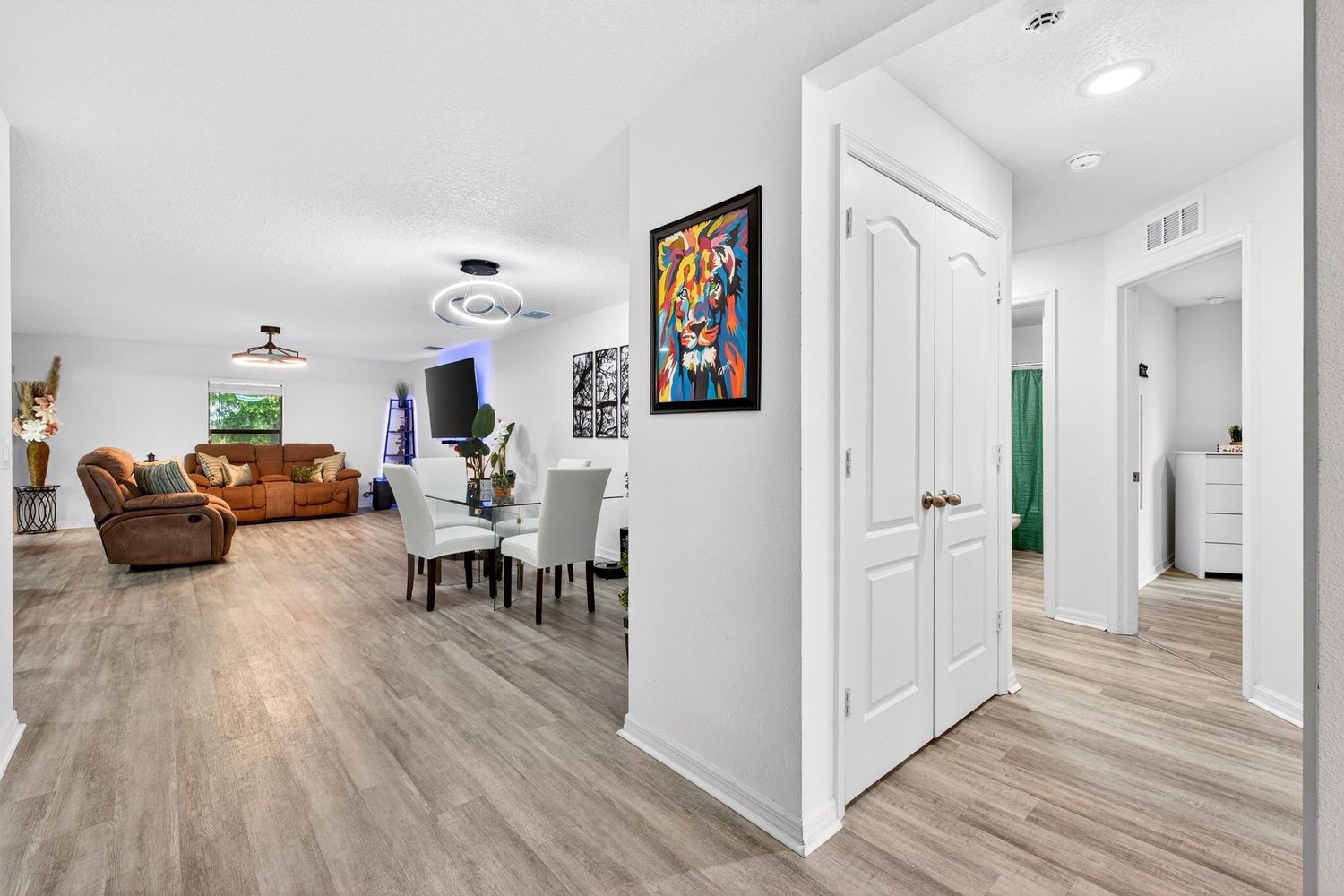 Bright open-concept living and dining area with light wood floors, white walls, ceiling fan, and colorful tiger artwork.