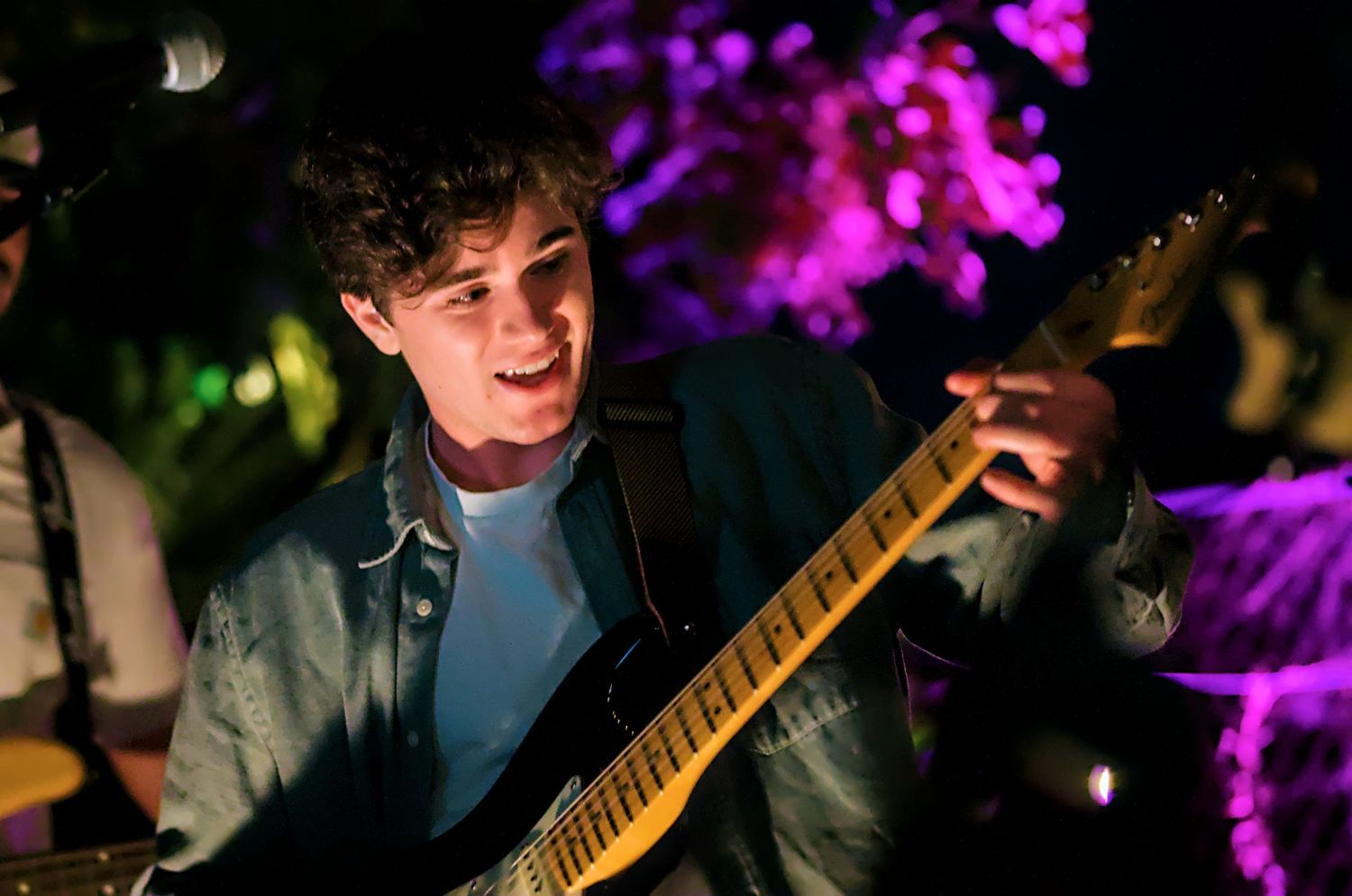 Guitarist performs on stage with purple and green lighting effects creating a dramatic concert atmosphere.