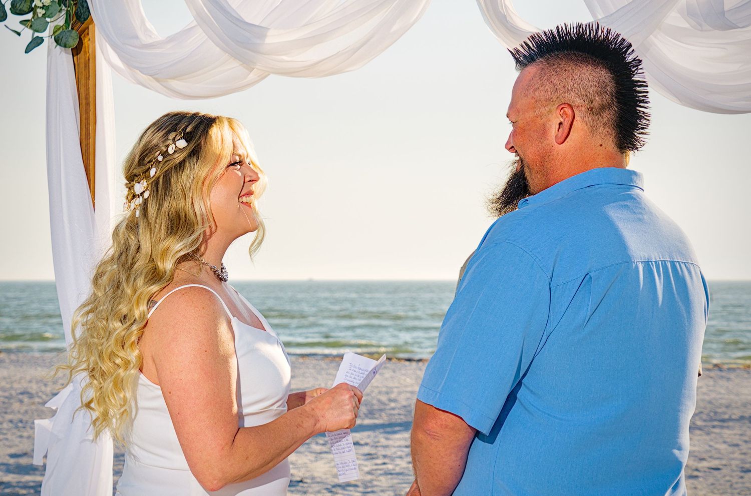 Couple shares romantic moments during a beach photoshoot with white curtains and greenery in the background.