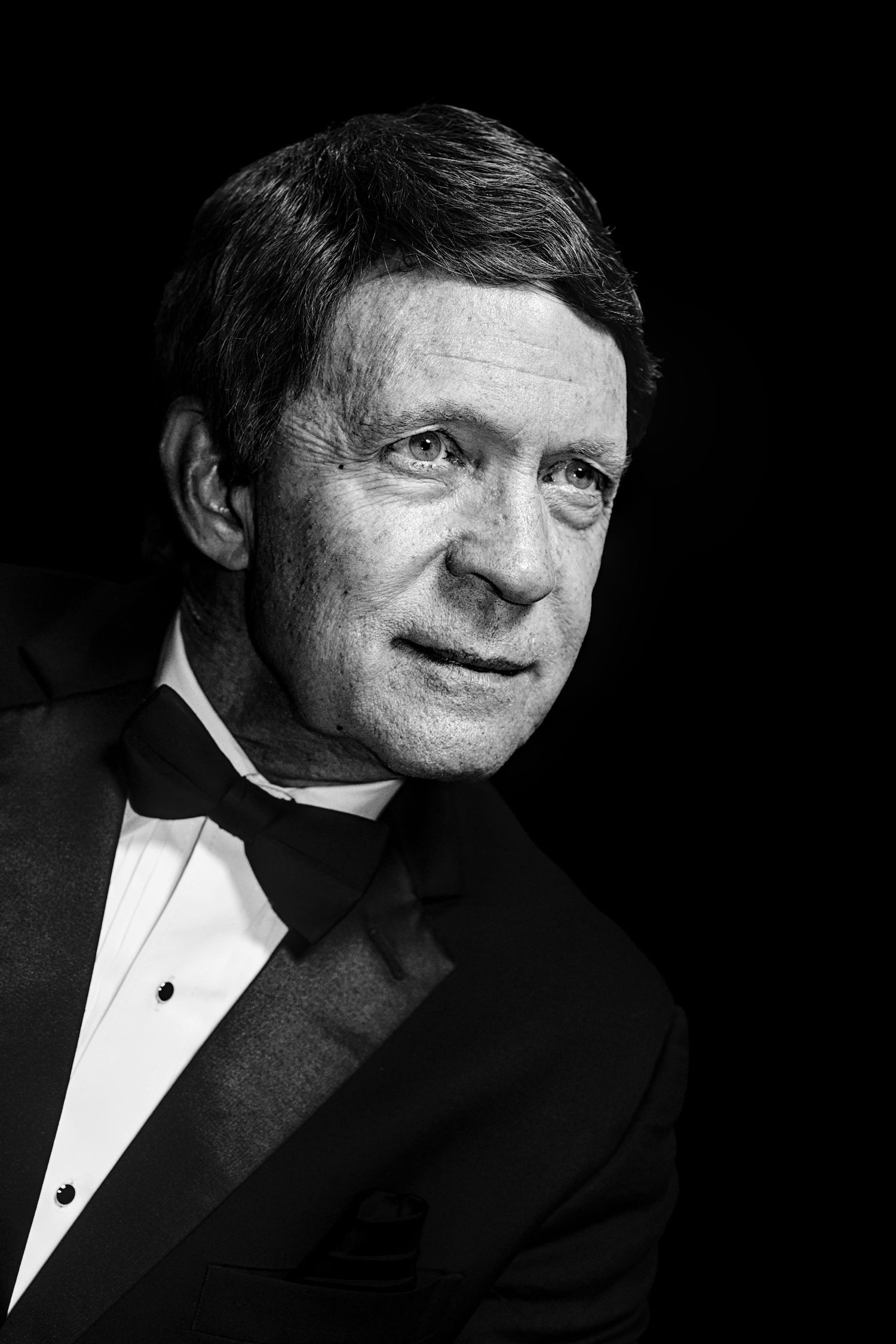 Black and white portrait of an elegant middle-aged man in a tuxedo with bow tie against dark background.