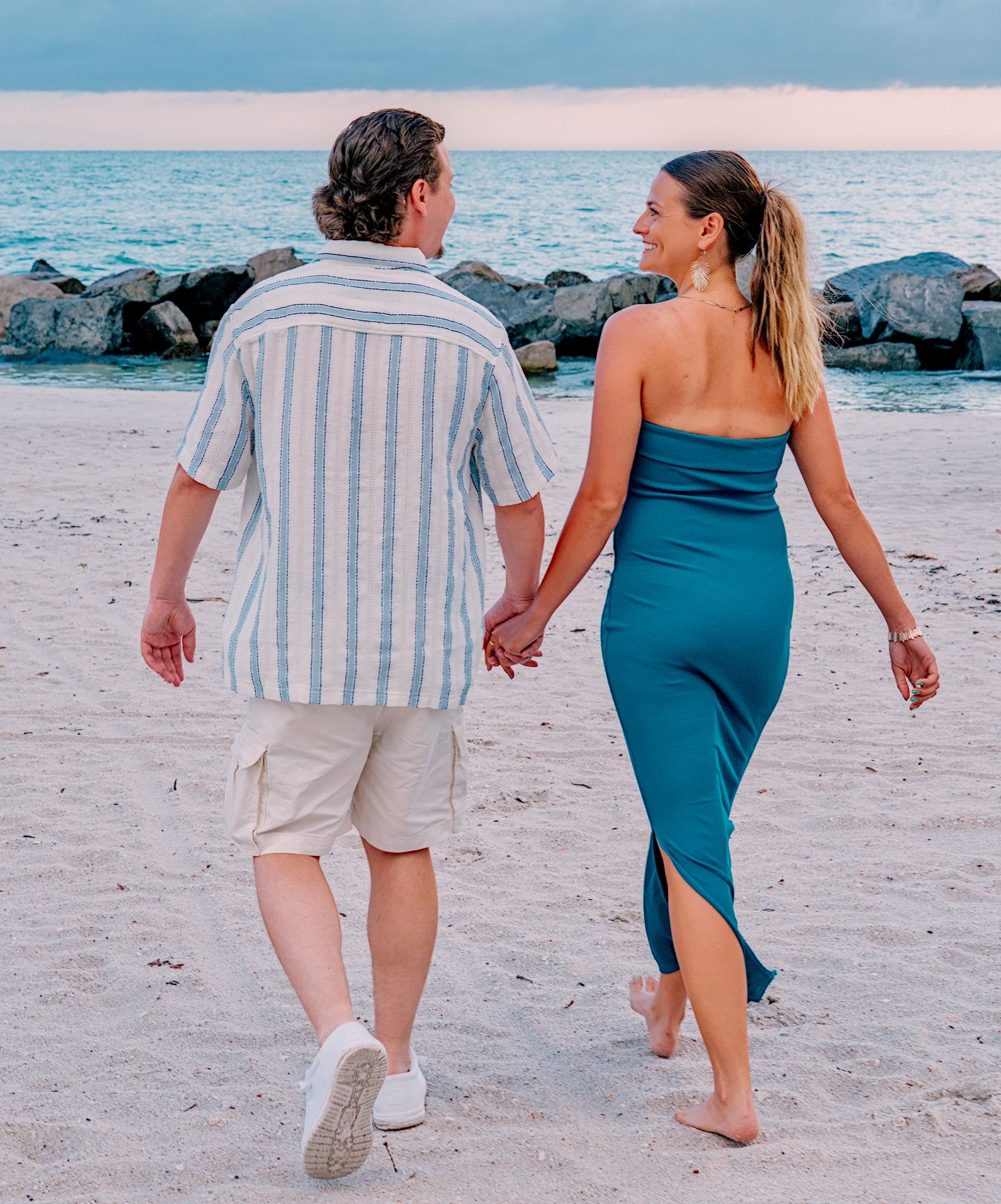 Couple holding hands walking on a sandy beach at sunset, man in striped shirt and woman in teal dress.