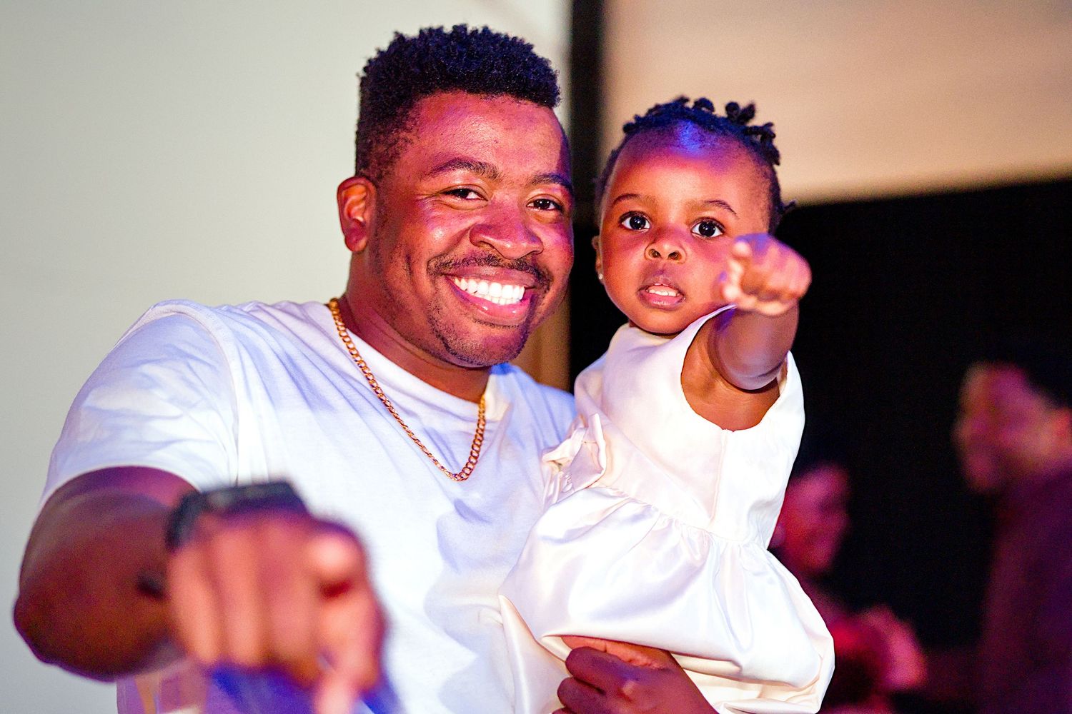 A smiling man in a white shirt holds a young toddler dressed in white while both point at the camera.