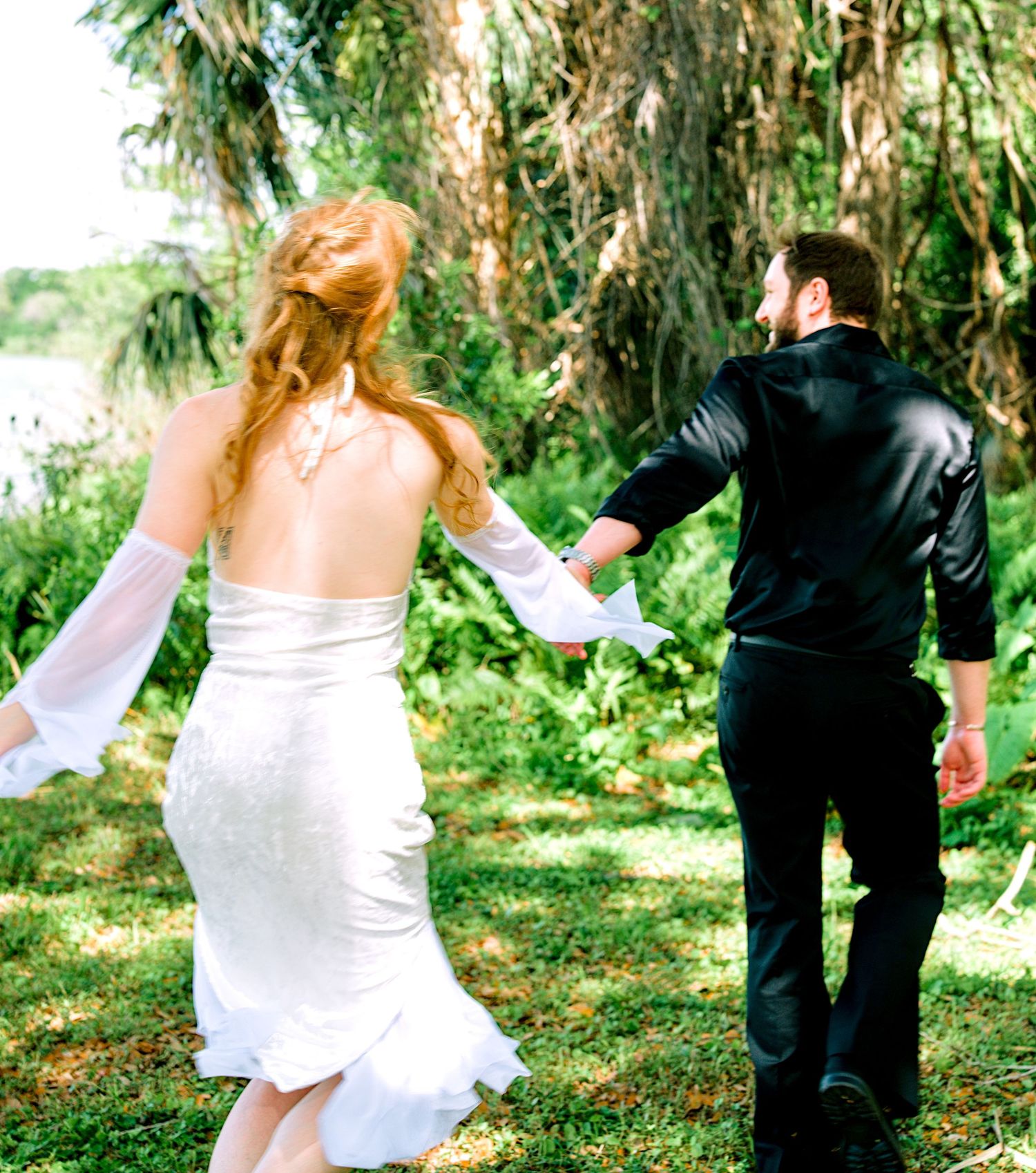 Bride in white backless gown holds groom's hand as they walk through lush green garden.