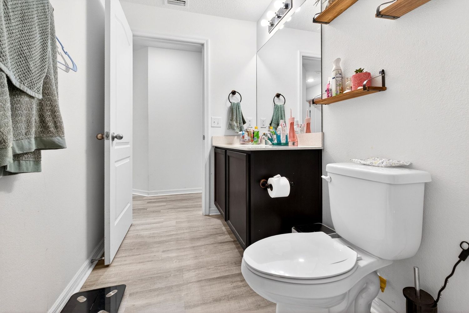 Modern bathroom with wood-look floors, dark vanity, white toilet, and open shelving with decor accents.