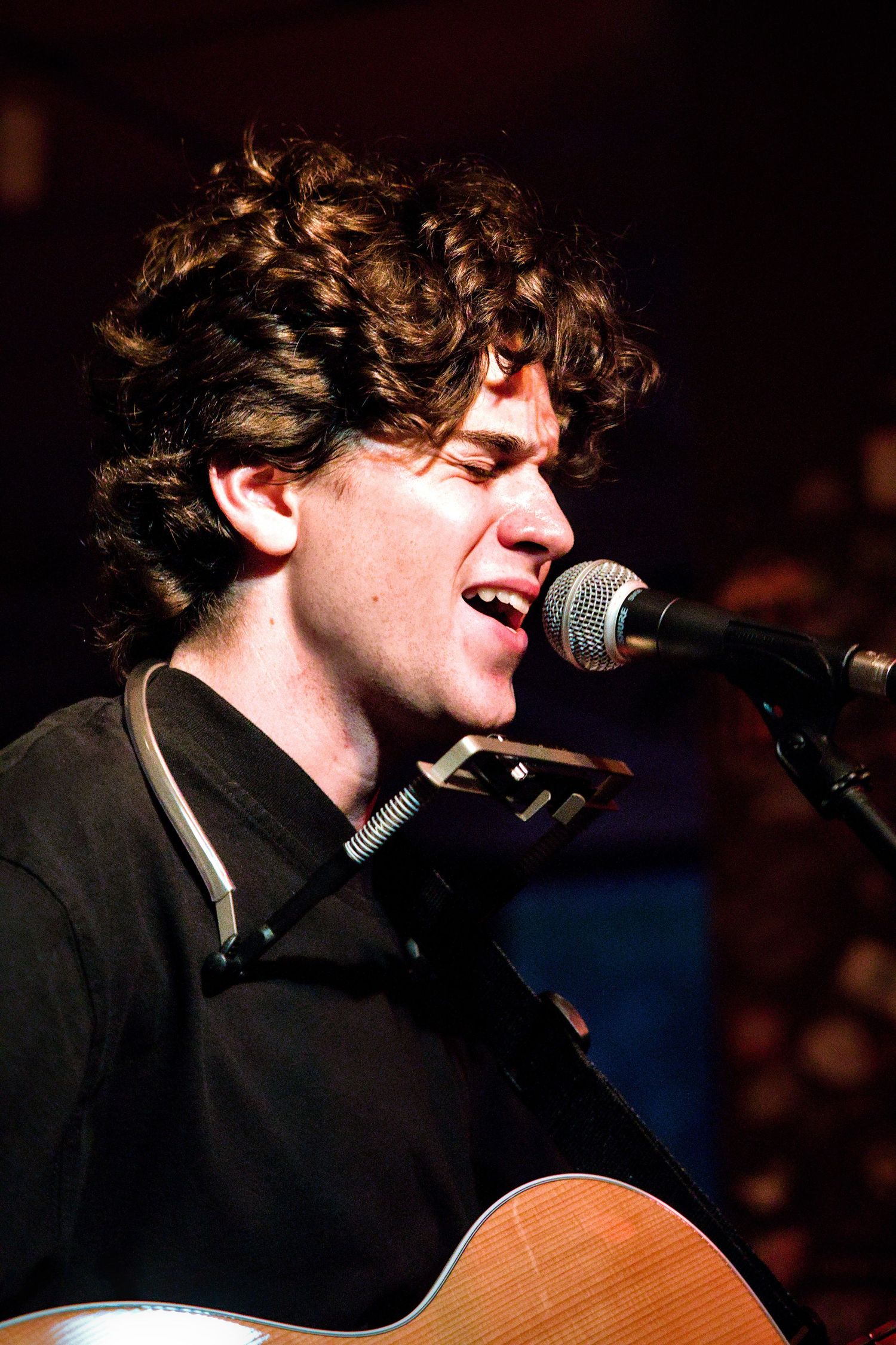 Young curly-haired male musician singing into microphone while playing acoustic guitar on stage.