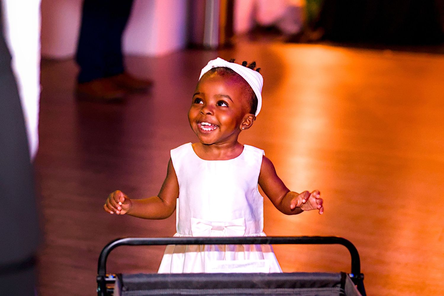 Happy toddler girl in white dress standing at a walker on a dance floor.