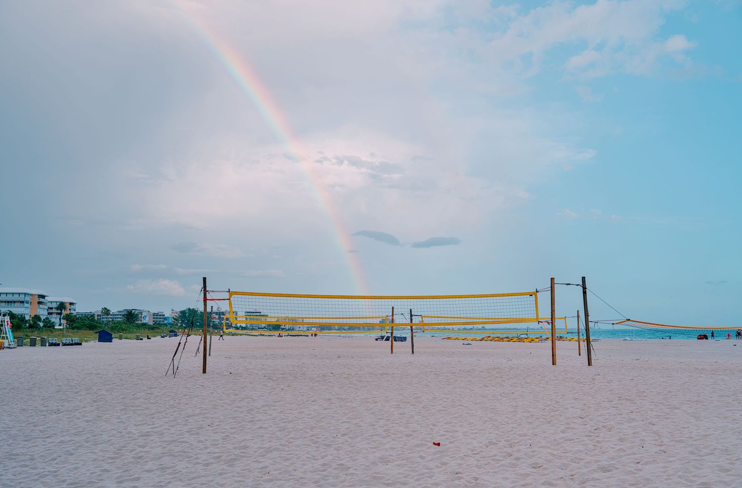 Beach volleyball nets on sandy shore with a rainbow arching over the cloudy blue sky in the background.