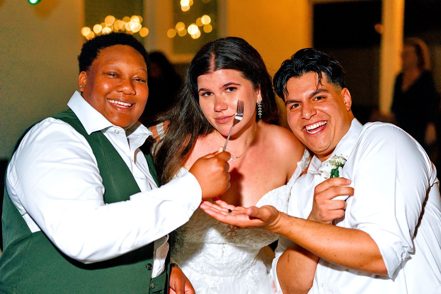 Two men and a bride smile while cutting wedding cake together at a reception.