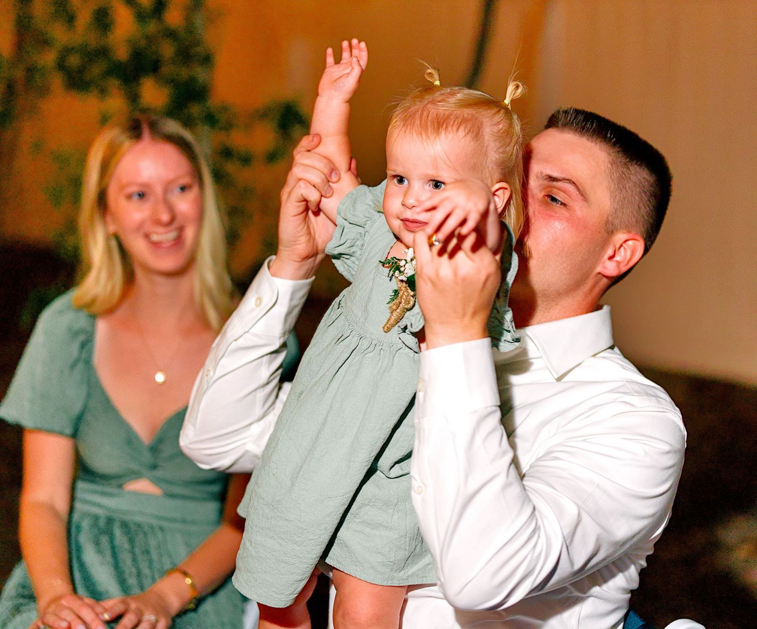 A man lifts a smiling toddler girl in the air at a wedding reception while a woman watches.