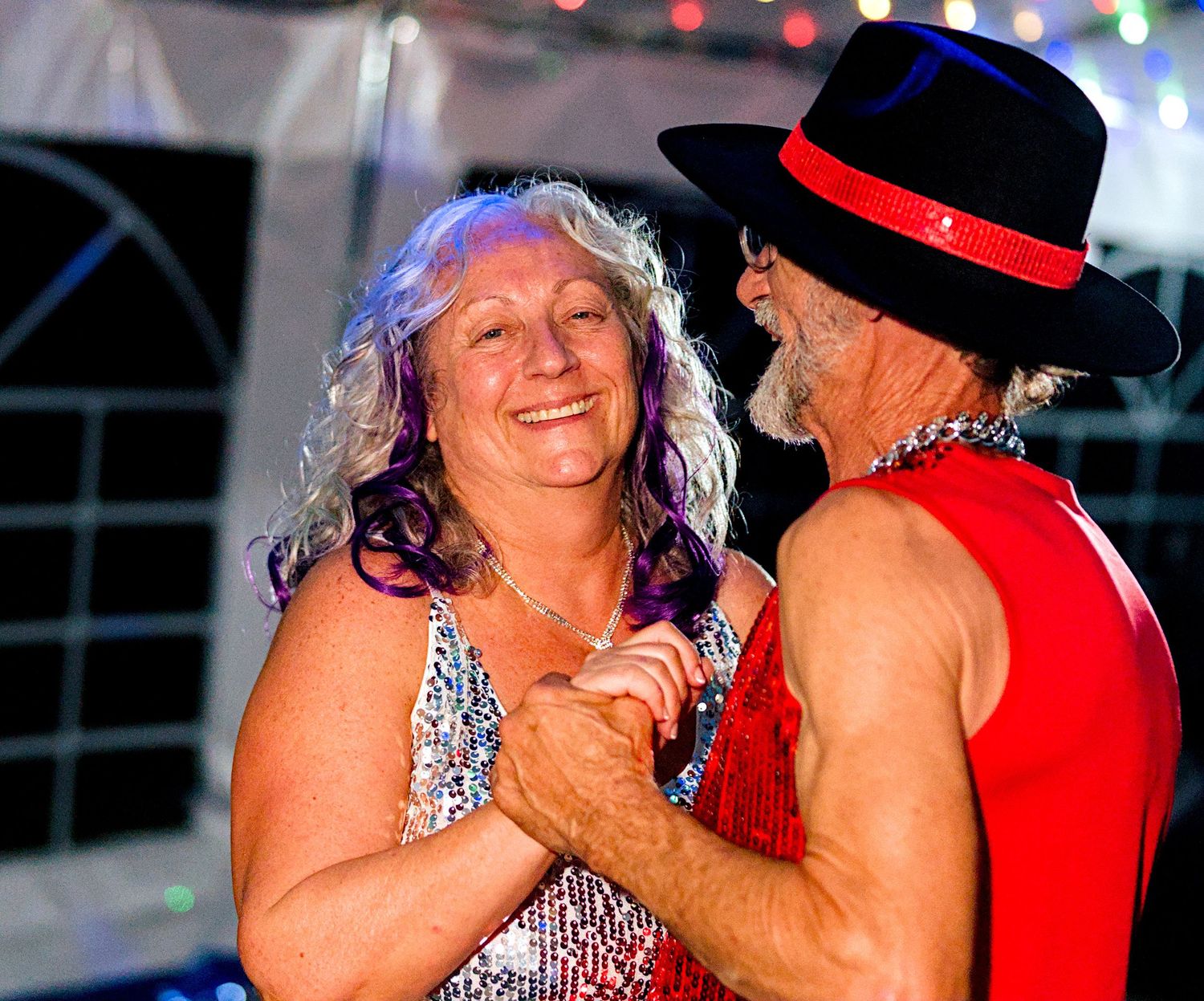 Older couple dancing together at a party, woman in sequin dress, man in red vest and black fedora hat.
