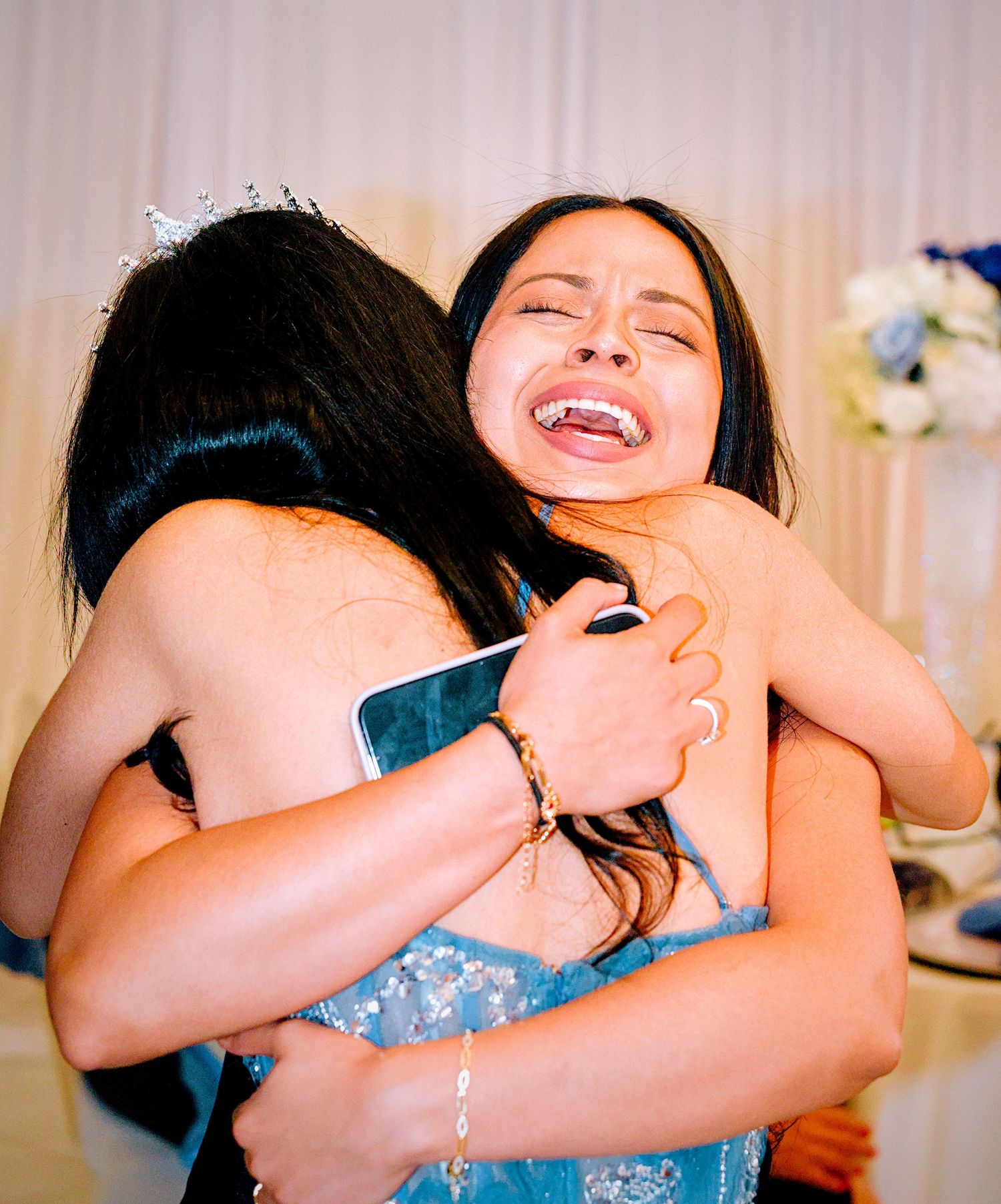 Two young women share a joyful embrace at a formal celebration, one wearing a tiara and blue dress.