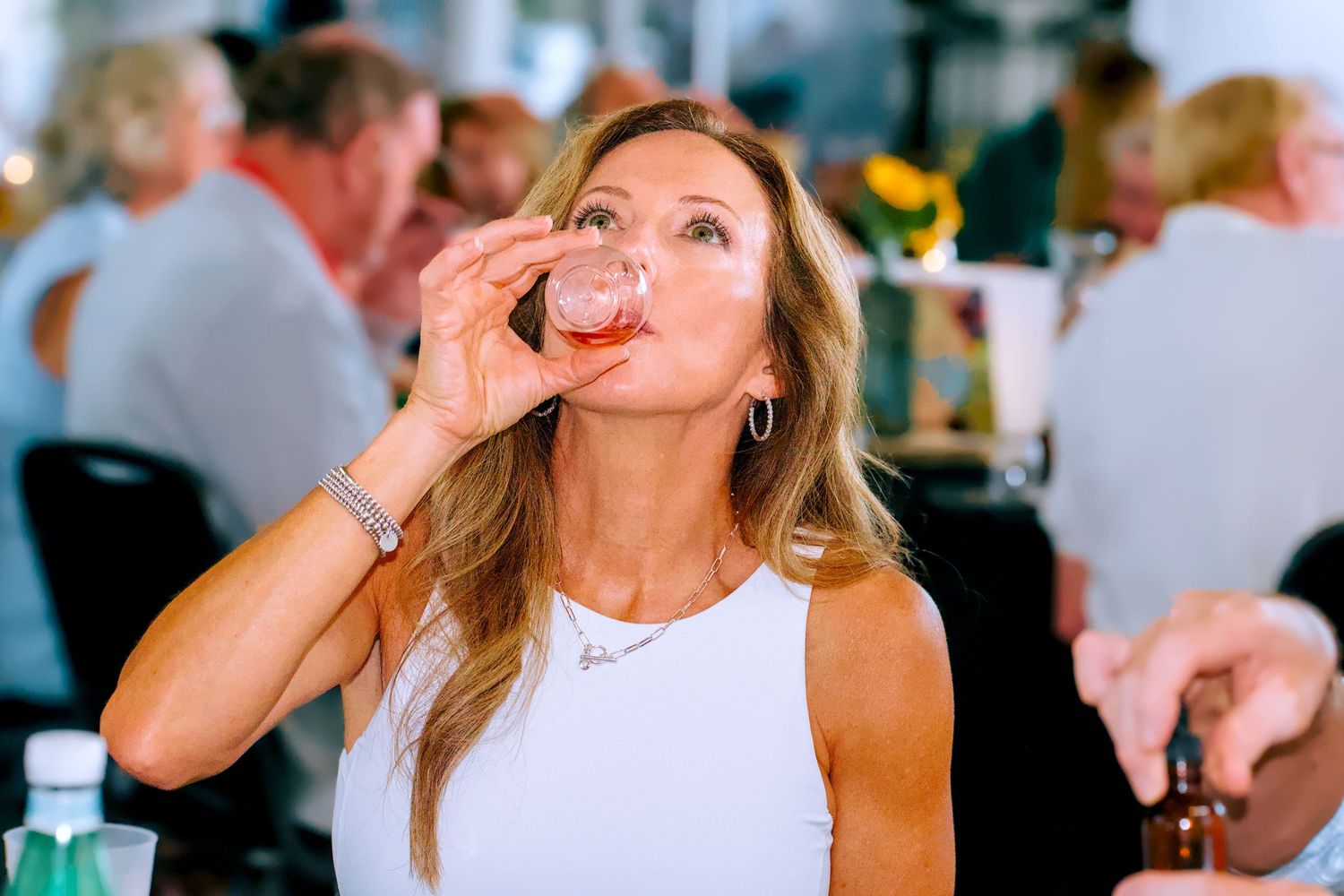 Woman in white top taking a shot drink at a social gathering event indoors.