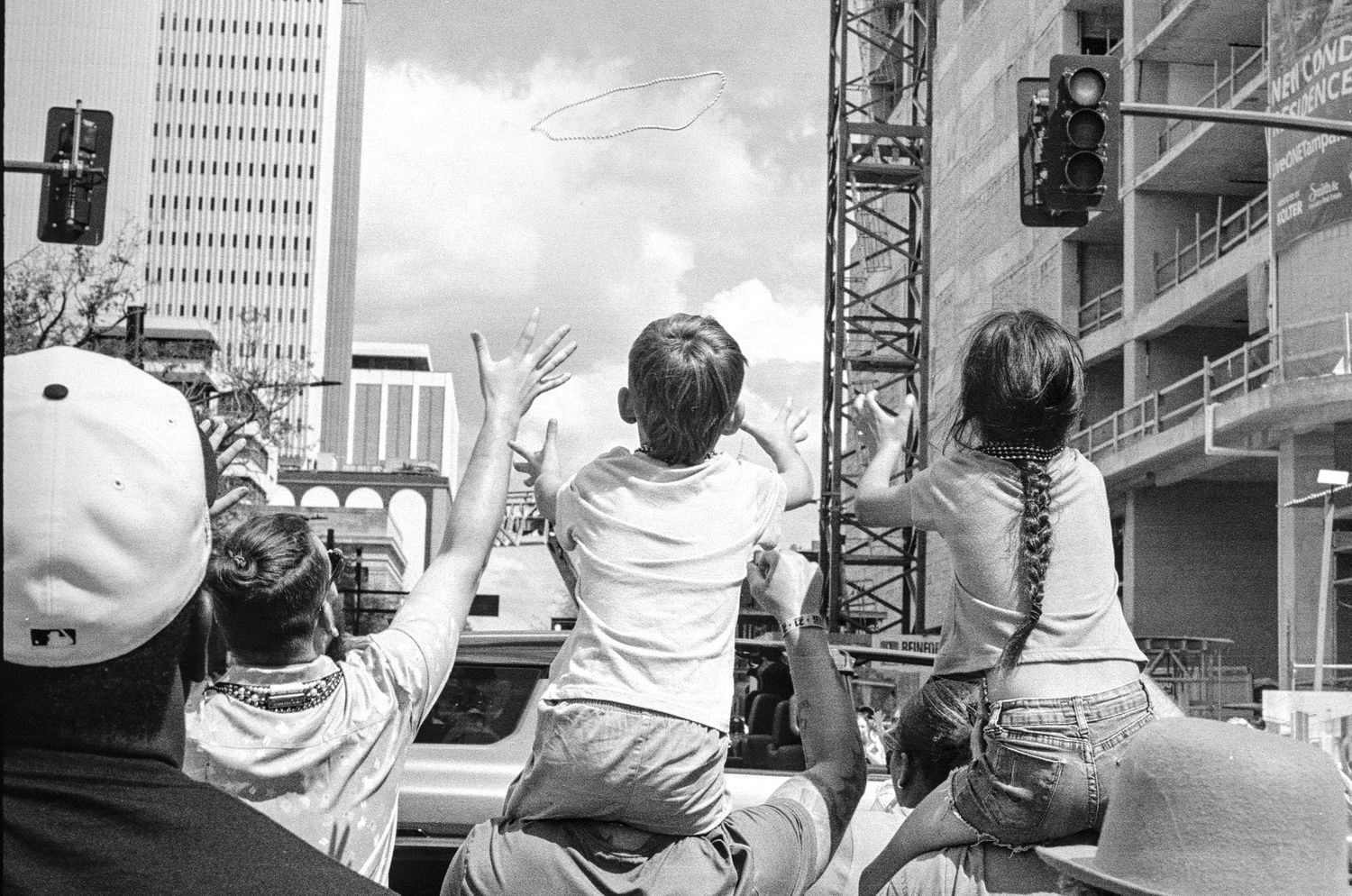 Crowd of people waving at a parade in an urban setting with construction cranes and city buildings in background.