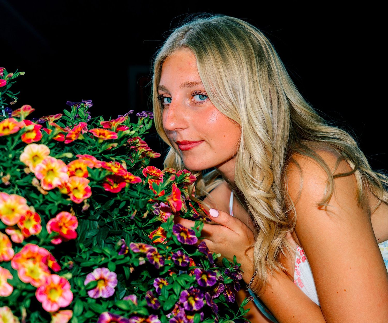 Young blonde woman smelling colorful orange and pink flowers in a garden at night.