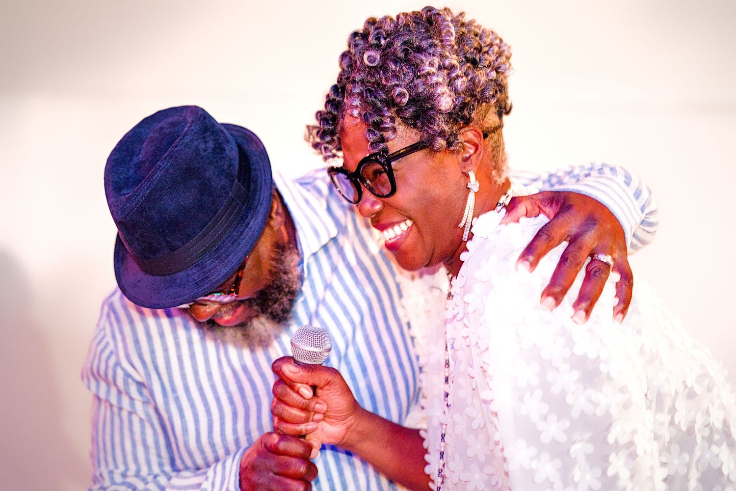Two joyful people laughing and embracing, one holding a microphone, sharing a warm moment together.