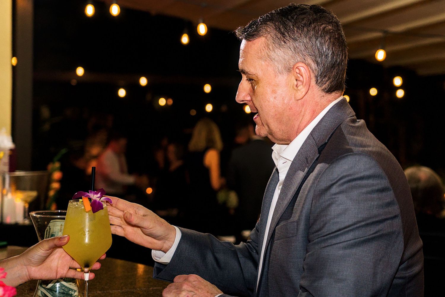 Man in suit smiling while receiving a garnished cocktail at an outdoor evening bar event with string lights.