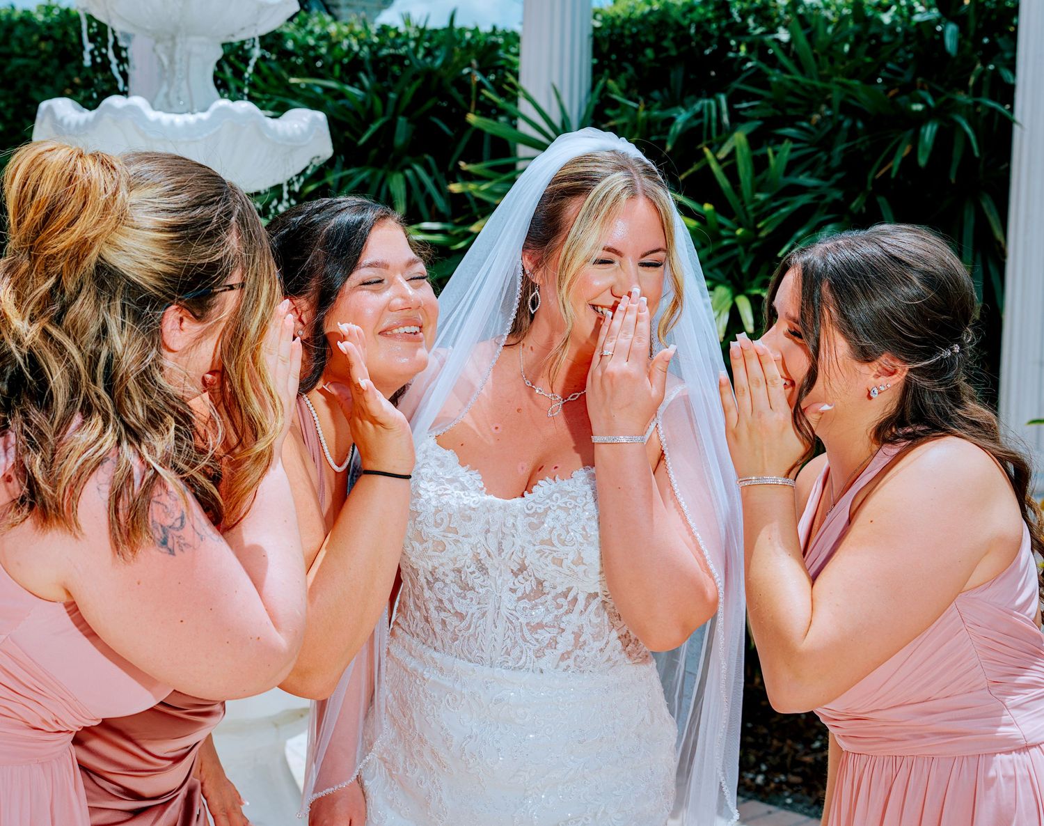 Bride in white lace gown and veil shares emotional moment with bridesmaids in blush pink dresses outdoors.