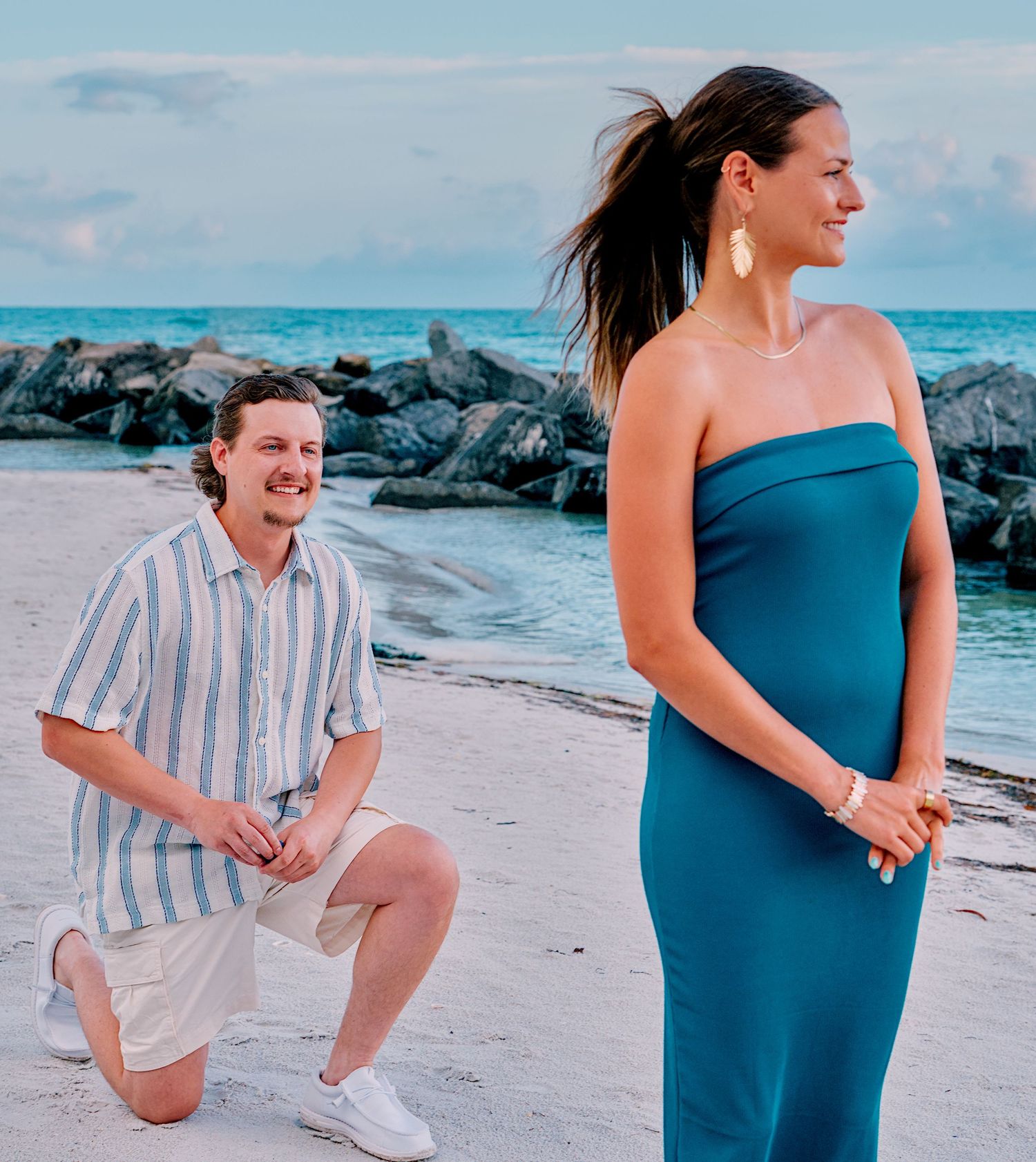 Man kneeling on beach proposing to woman in teal strapless dress near rocky shoreline under blue sky.
