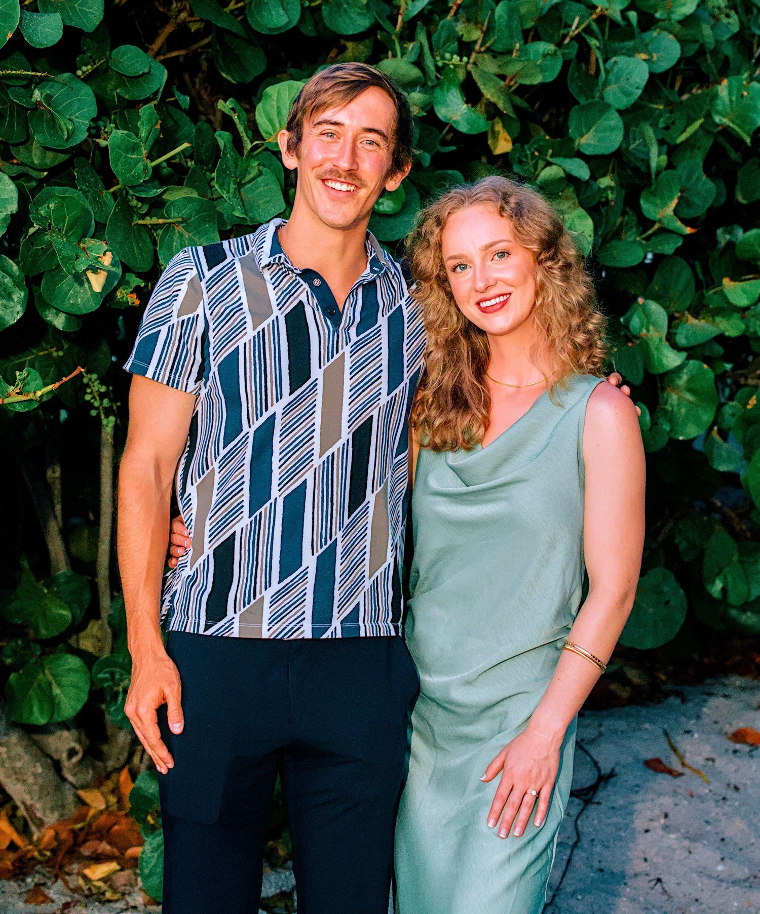 A smiling couple poses outdoors in front of lush green foliage, wearing coordinating blue and green outfits.