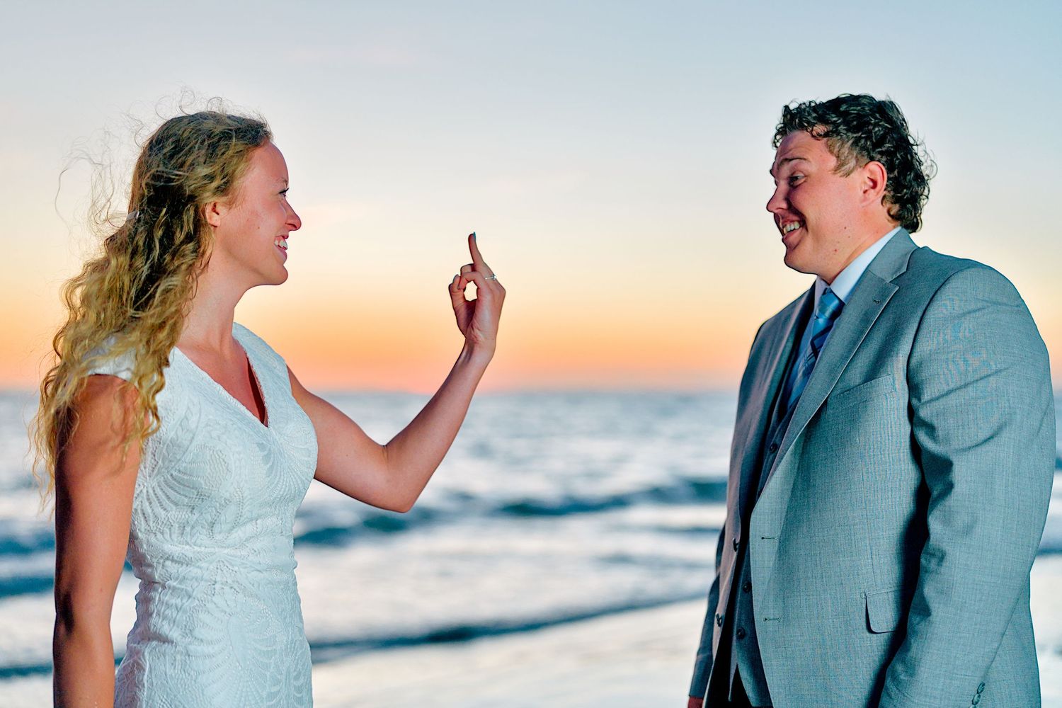 Bride in white dress playfully gesturing to laughing groom on beach at sunset.