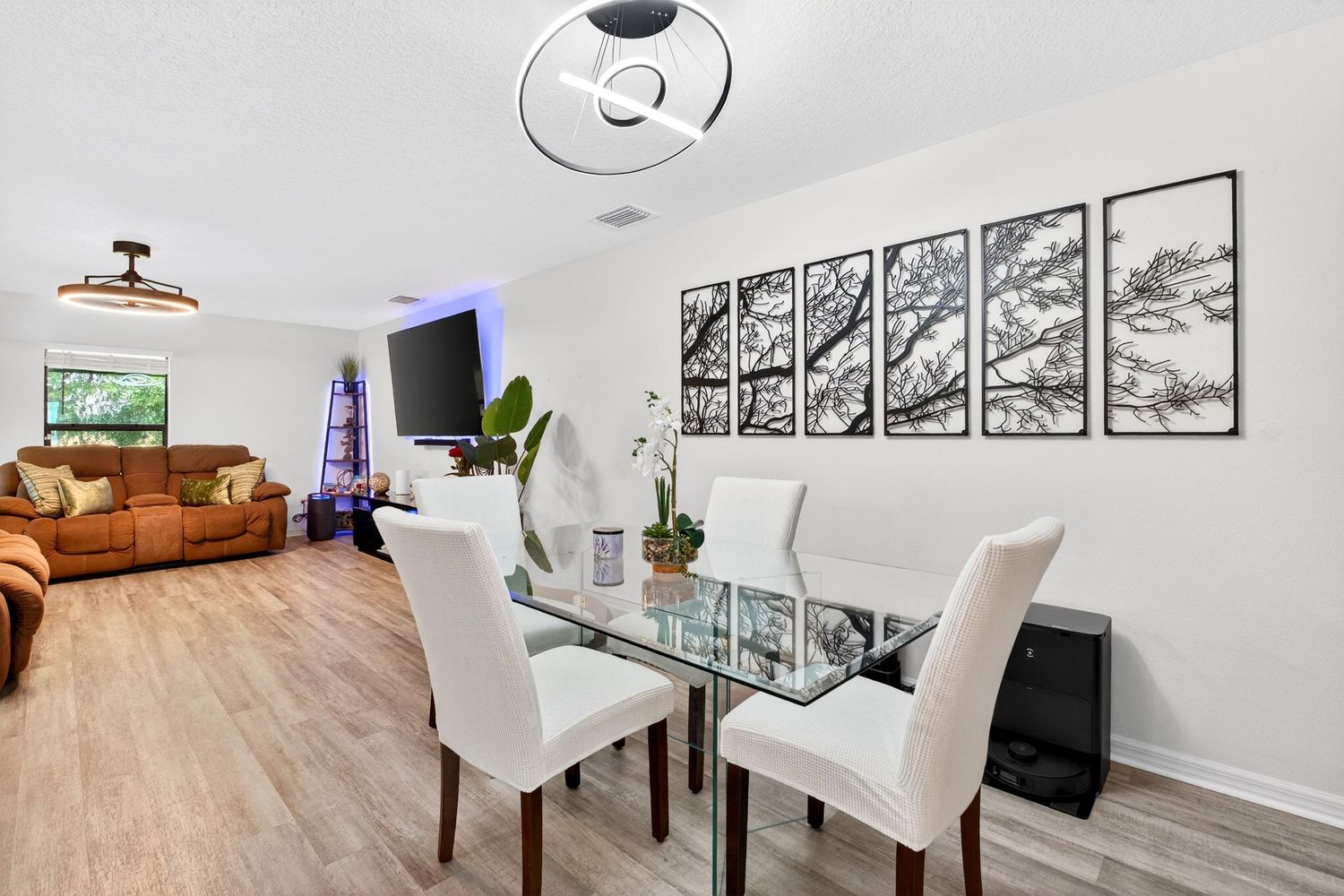 Modern dining area with glass table, white chairs, and tree wall art, open to a cozy living room with hardwood floors.